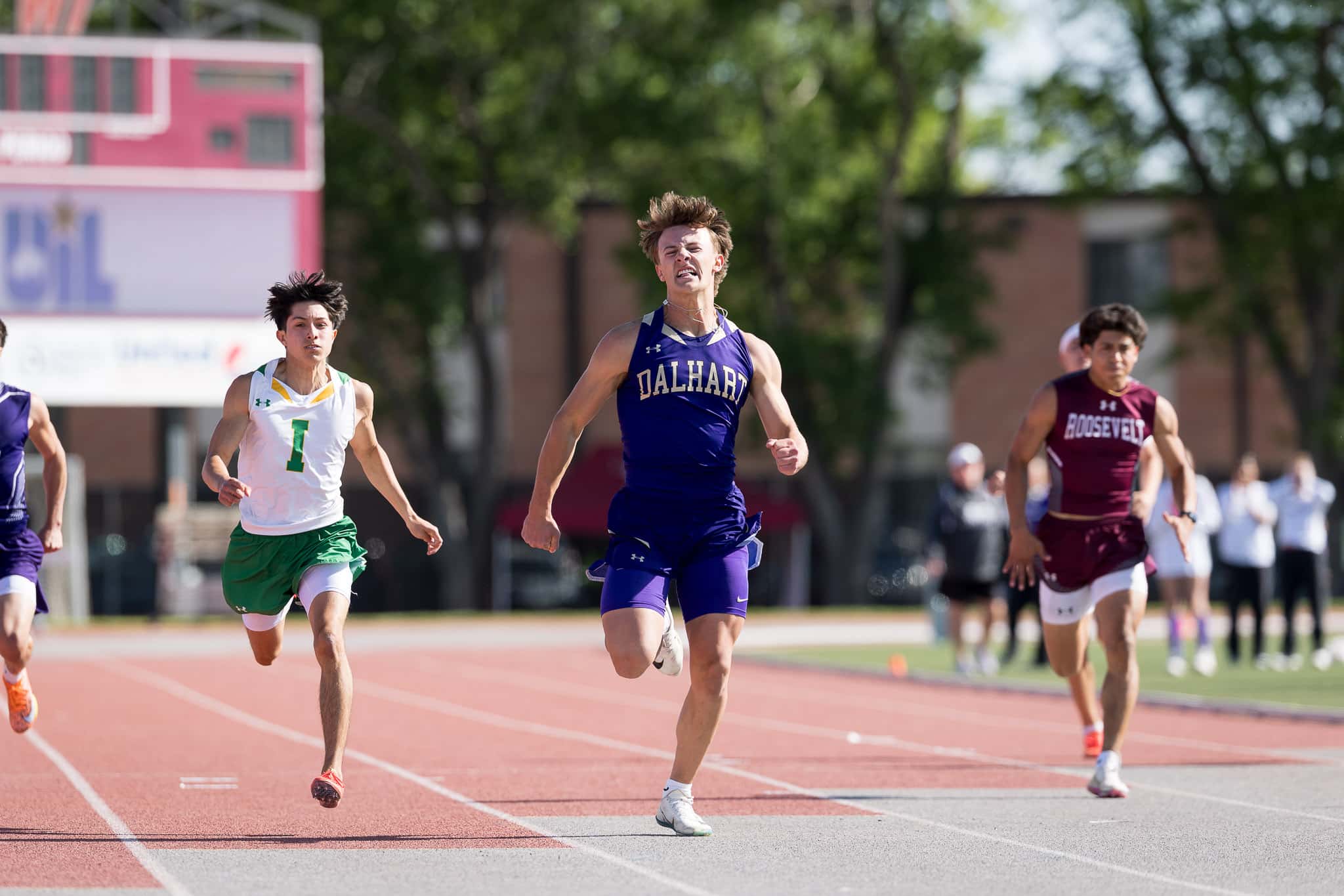 Male sprinters race on a red track; center athlete in a purple DALHART uniform leads flanked by competitors in white/green and maroon.