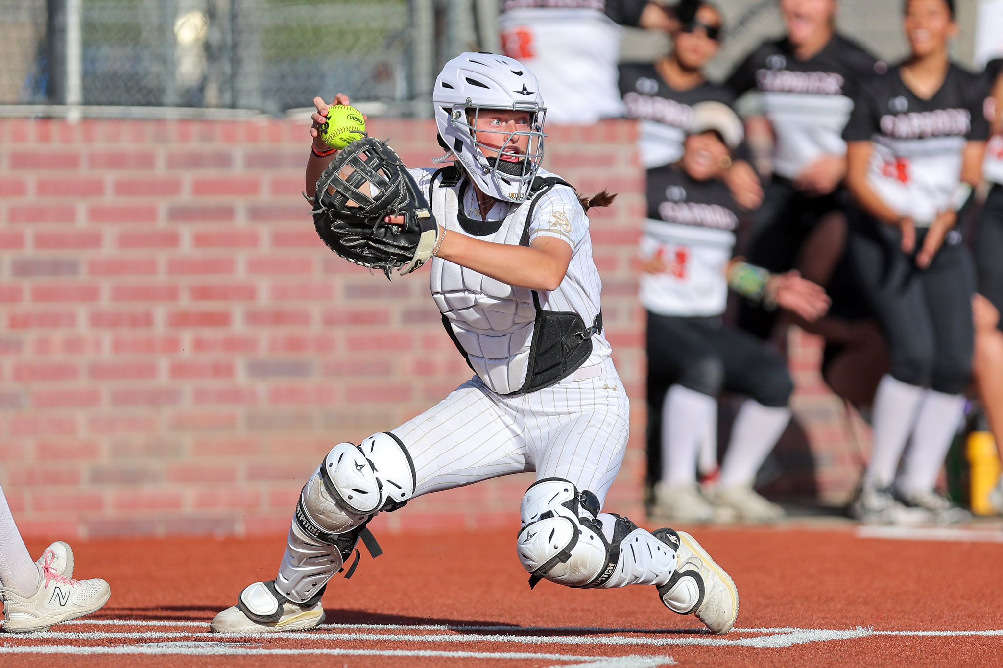 Softball catcher in white pinstriped gear and helmet running on the field, with a brick wall and cheering spectators in the background.