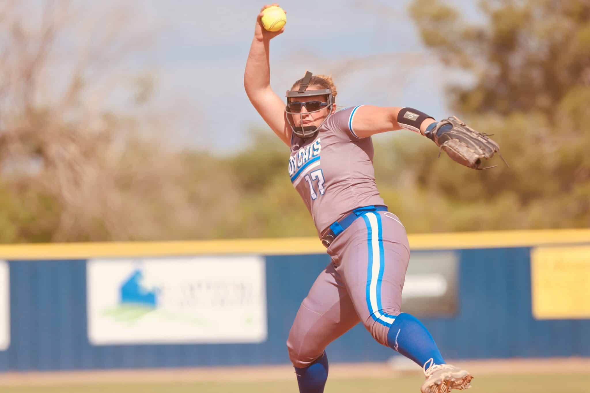 Female softball pitcher mid-throw in a gray-pink uniform with face guard, sunglasses, and glove, on a sunny field.