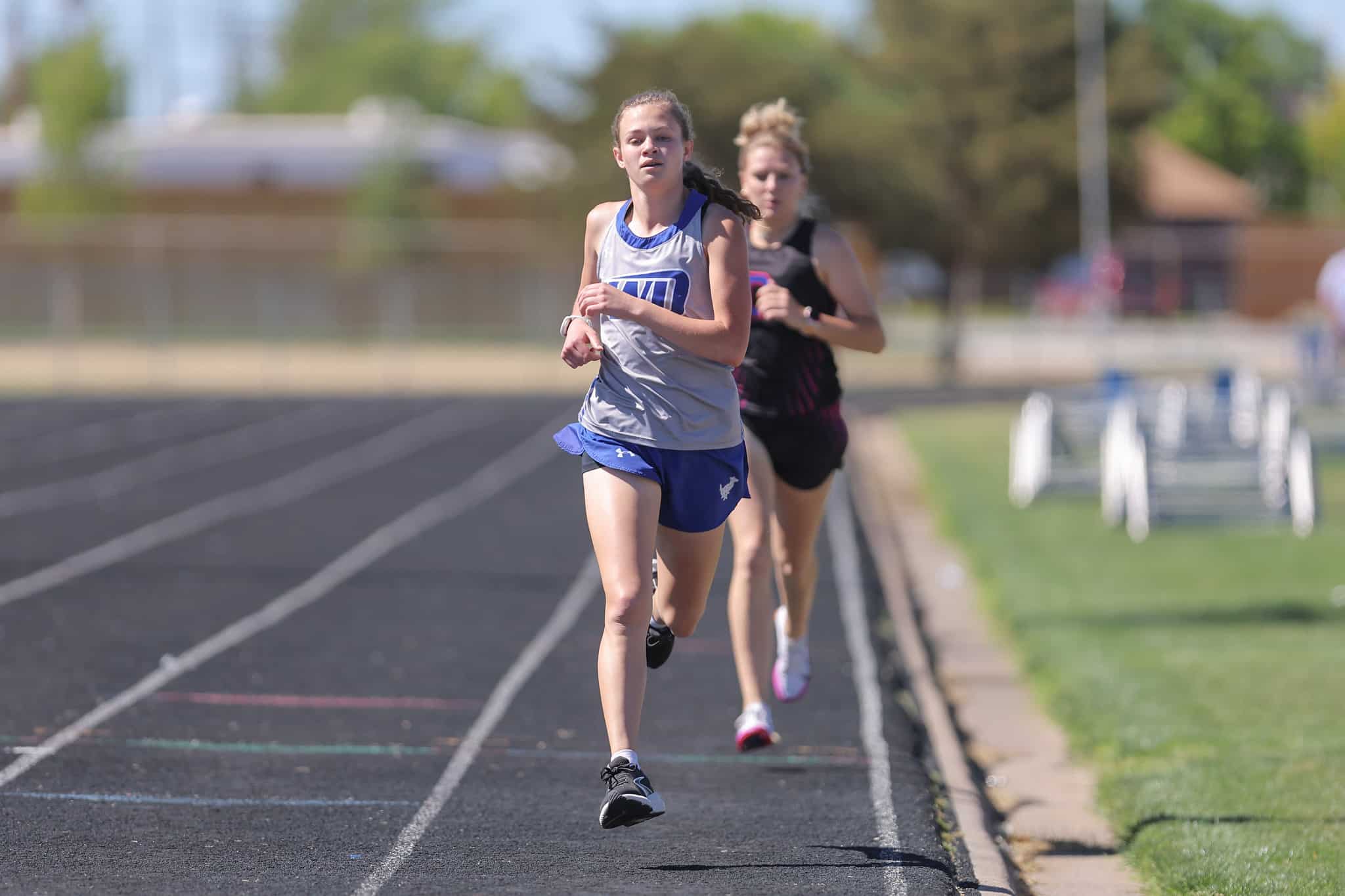 Female runner in gray and blue sprinting on outdoor track, leading a group of competitors behind her.