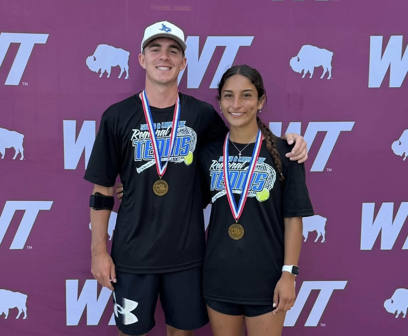 Two athletes in matching black shirts pose with gold medals around their necks in front of a purple WT backdrop with white logos and bison icons.