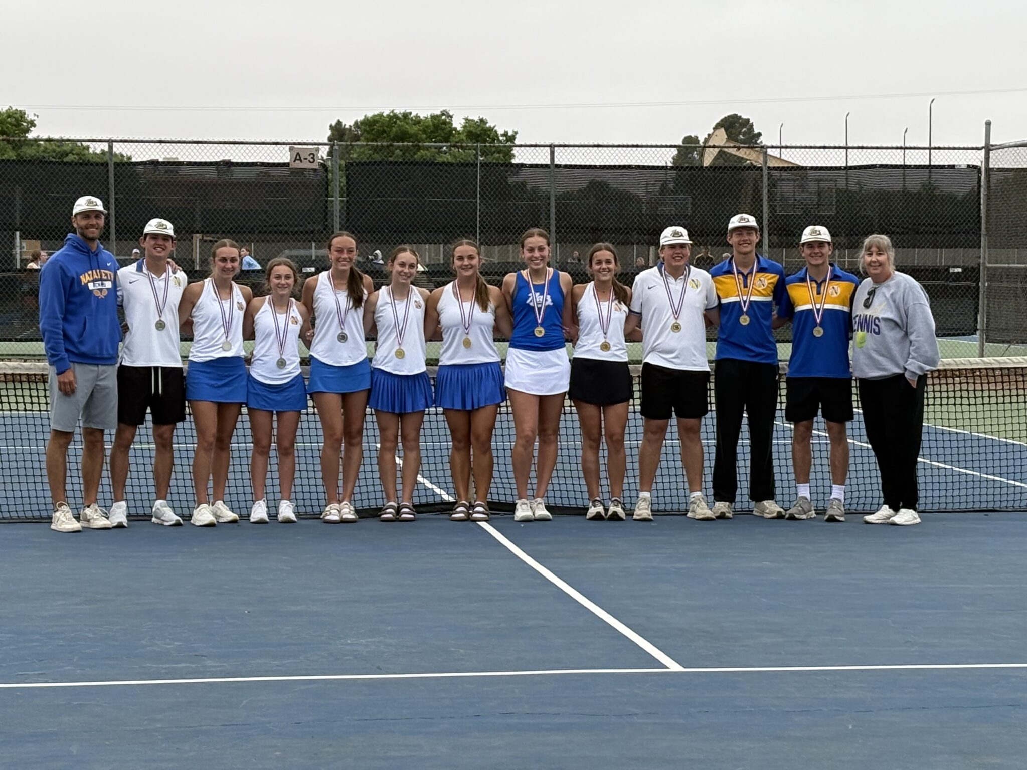 Group of young tennis players standing in a line on a blue court, wearing medals and white uniforms, with two coaches at each end, behind a chain-link fence after a match.