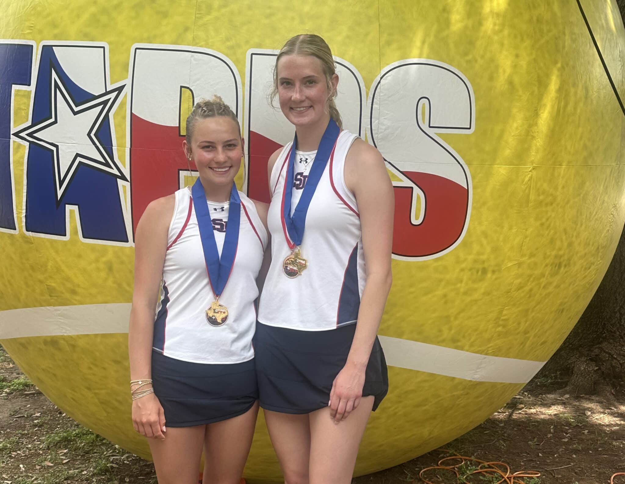 Two young female athletes wearing medals pose in front of a large yellow inflatable backdrop with a star logo on it.