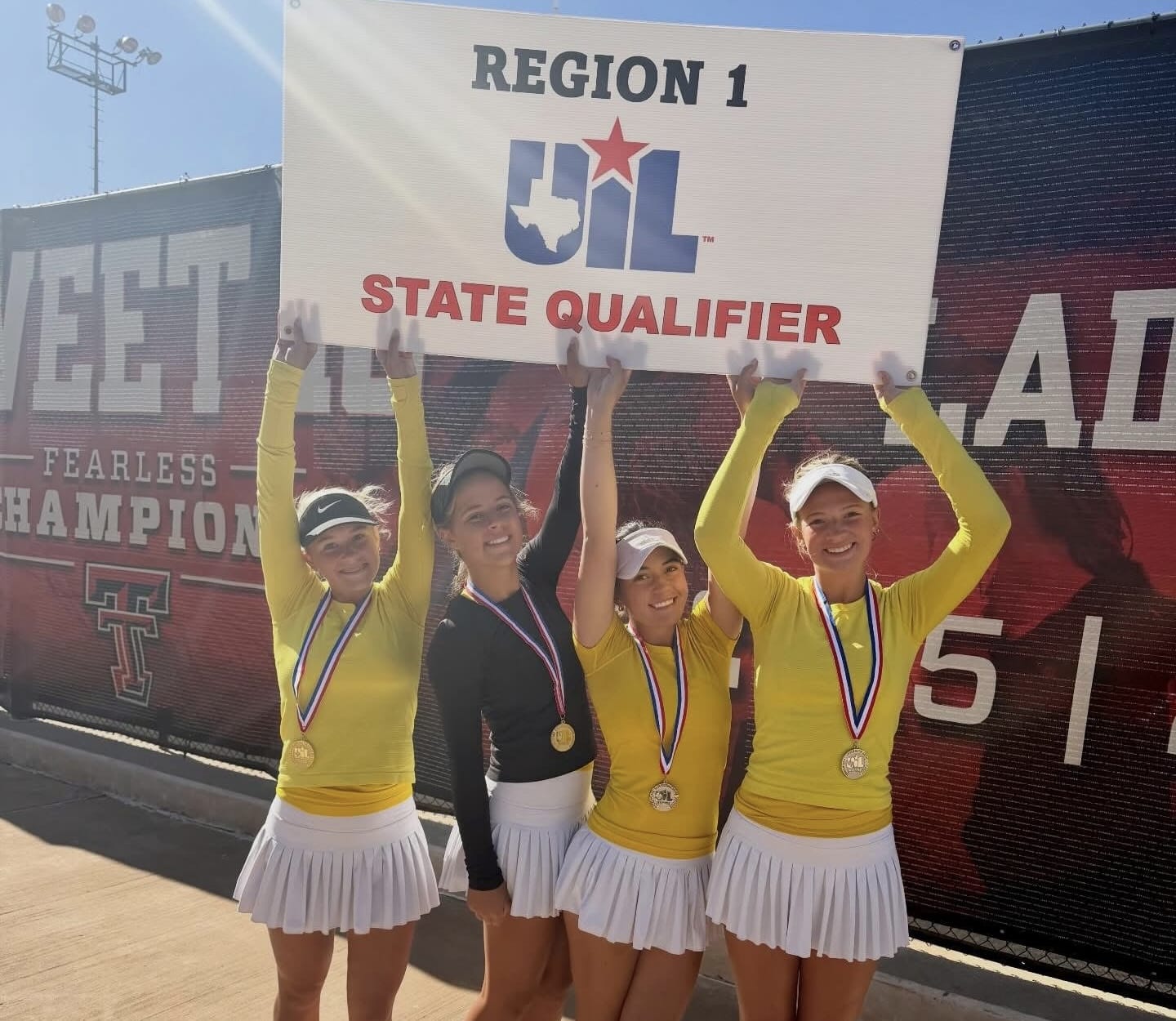 IMG_4837 | Press Pass Sports Four young female athletes in yellow tops and white skirts raise a UIL State Qualifier banner, wearing gold medals after a competition win.