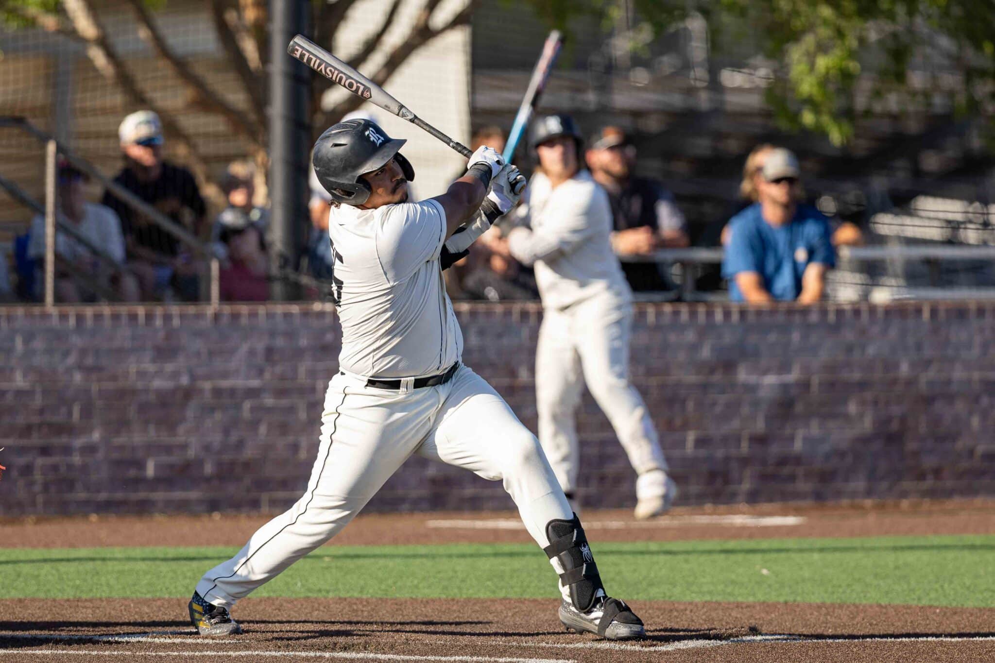 Baseball batter in white uniform swings at a pitch on a dirt infield with spectators in the background.