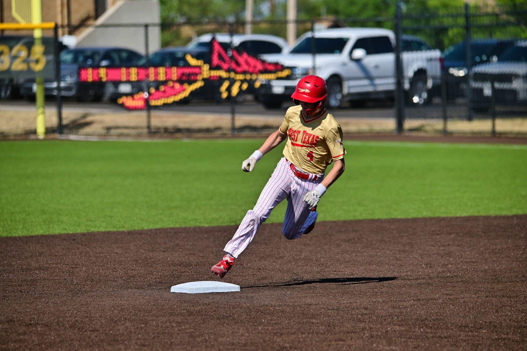 Baseball runner slides toward a white bag on infield dirt with a green outfield behind them, wearing a tan West Texas jersey and red helmet.