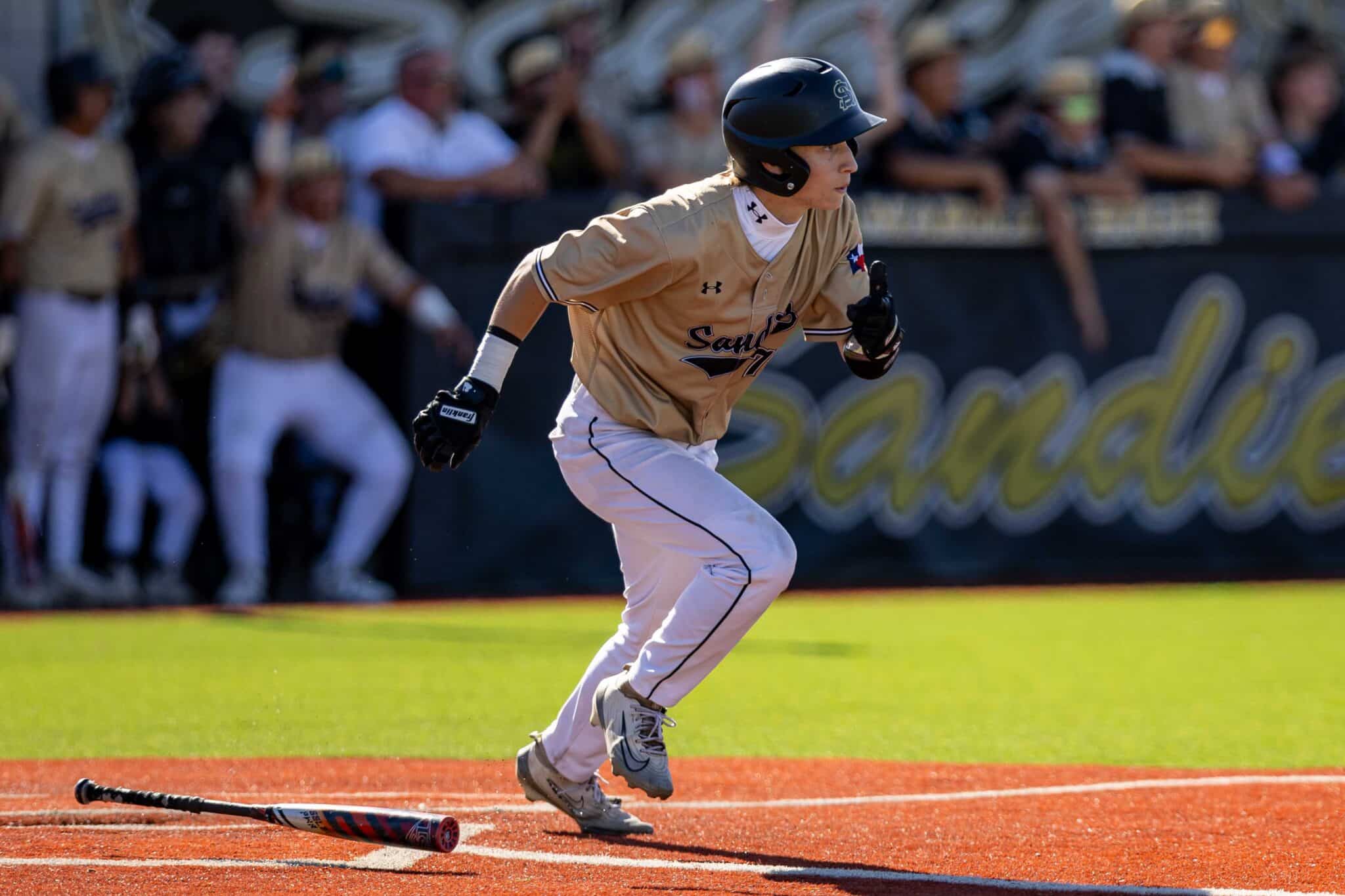 Baseball player in a tan uniform and helmet sprinting on the orange infield with a bat lying on the ground behind him and spectators in the background.