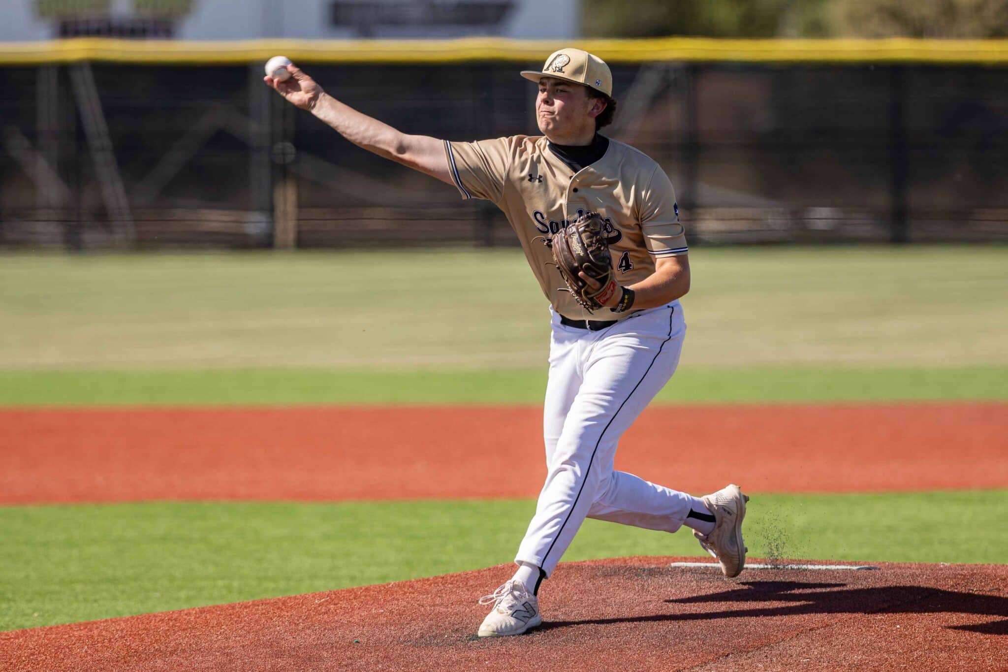 Baseball pitcher in a tan uniform winds up to throw on the mound, dirt in foreground and green outfield behind him.