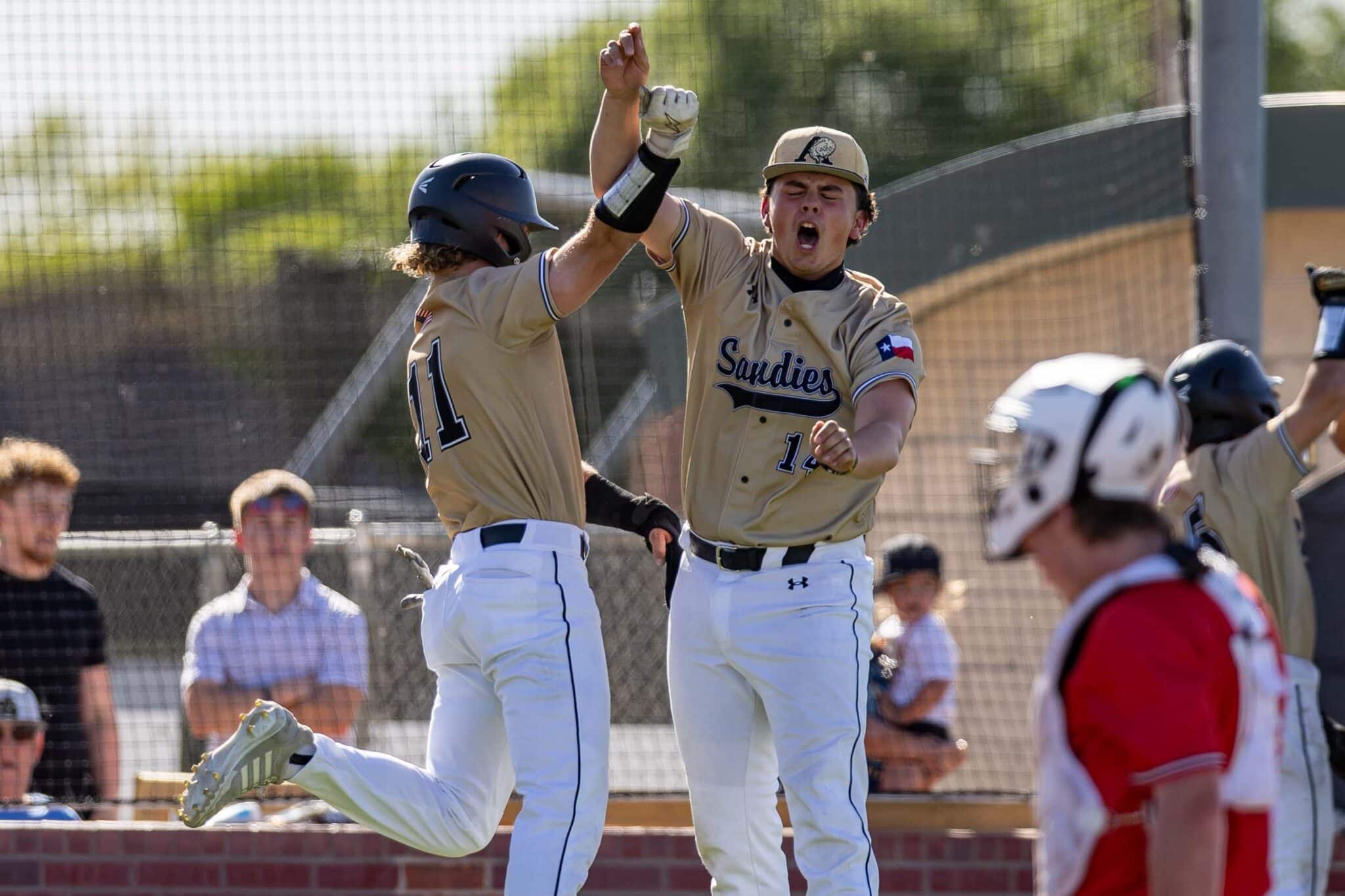 Two baseball players in tan uniforms celebrate with a high-five on the field as teammates applaud nearby behind a chain-link fence.