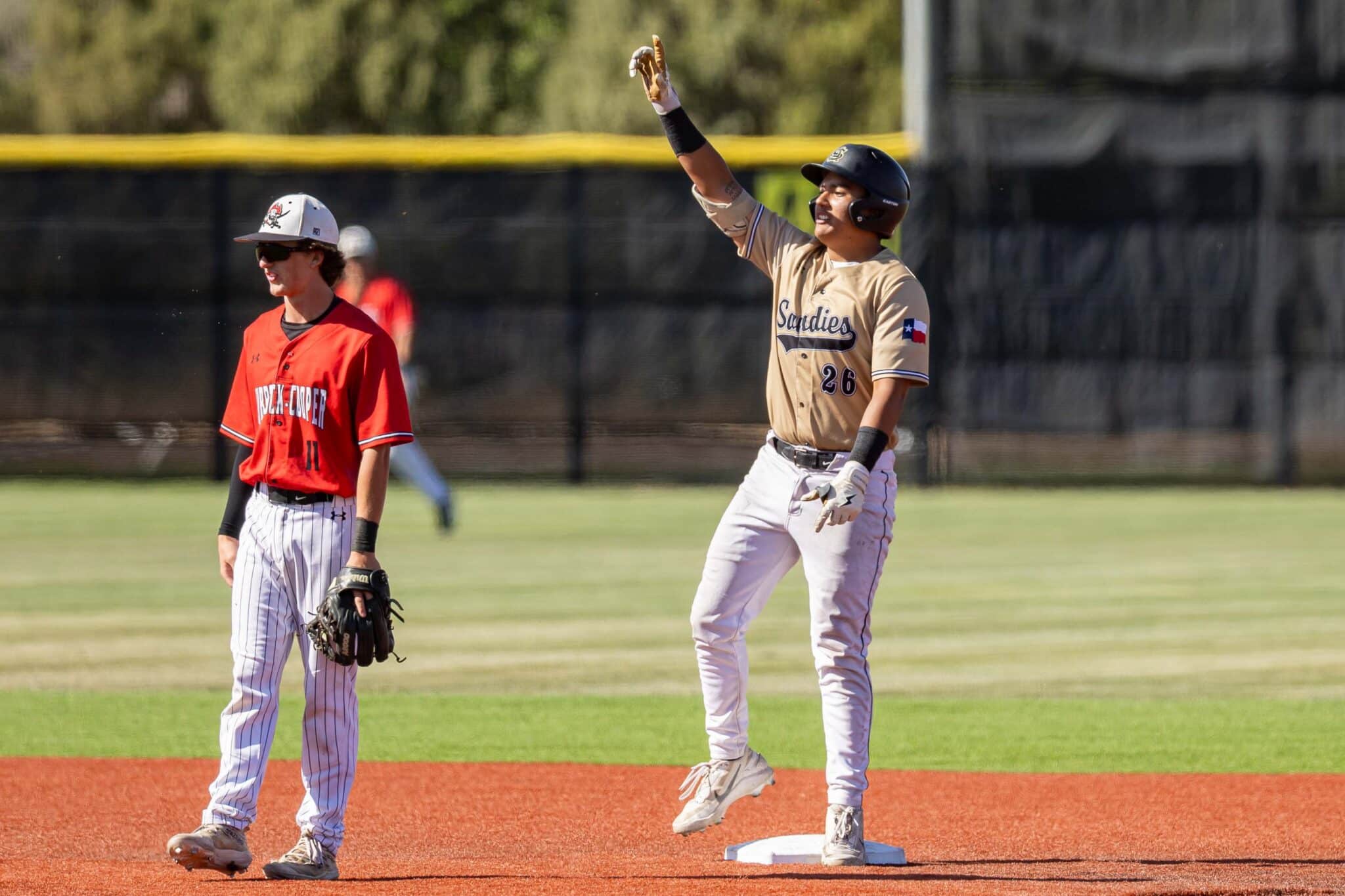 Two baseball players standing on a basepath; one in tan uniform raises his gloved hand in celebration while the other in red watches nearby.