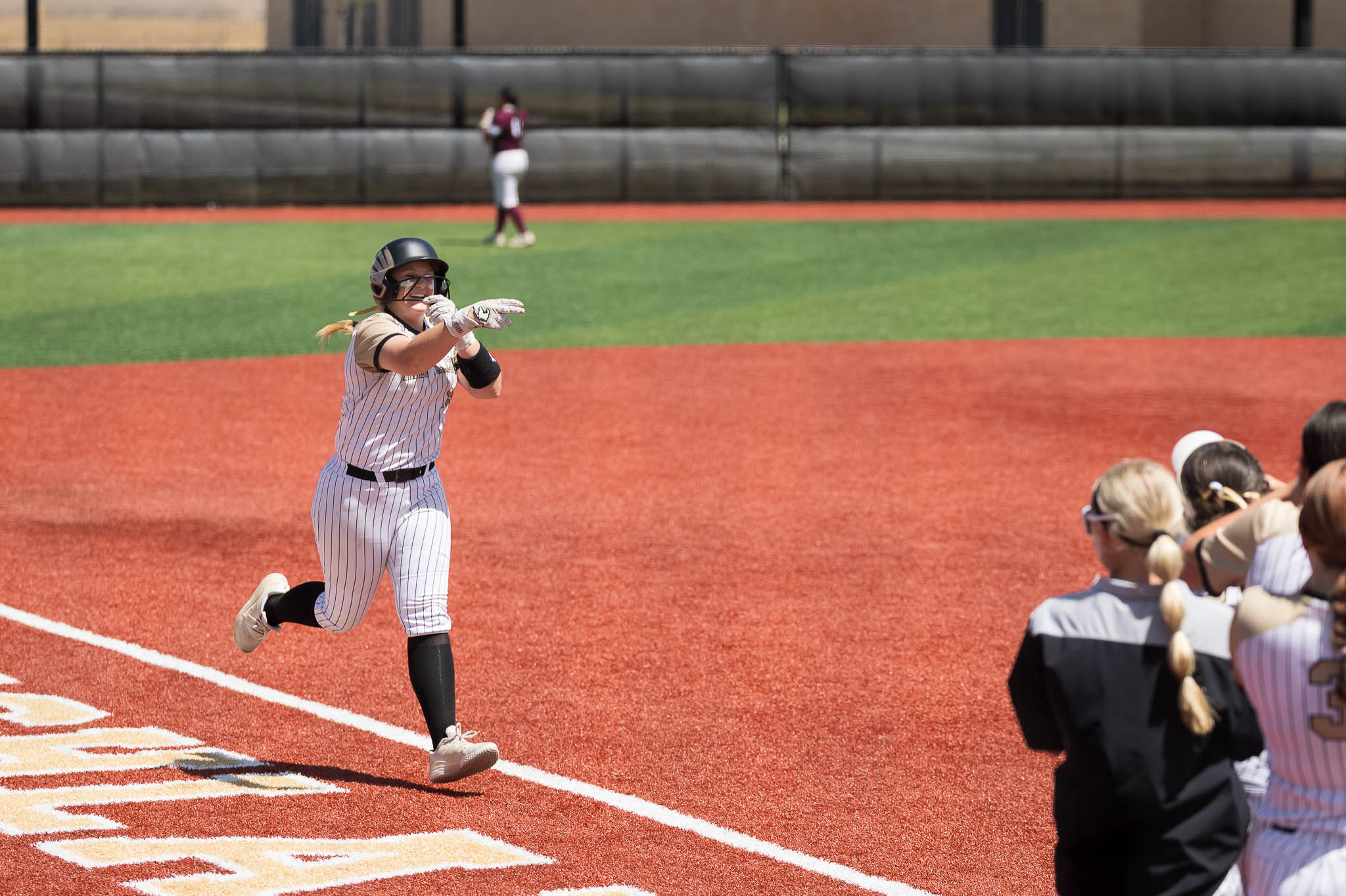 Softball player in a pinstriped uniform and helmet runs toward home plate on a red dirt infield, with teammates and spectators in the foreground.