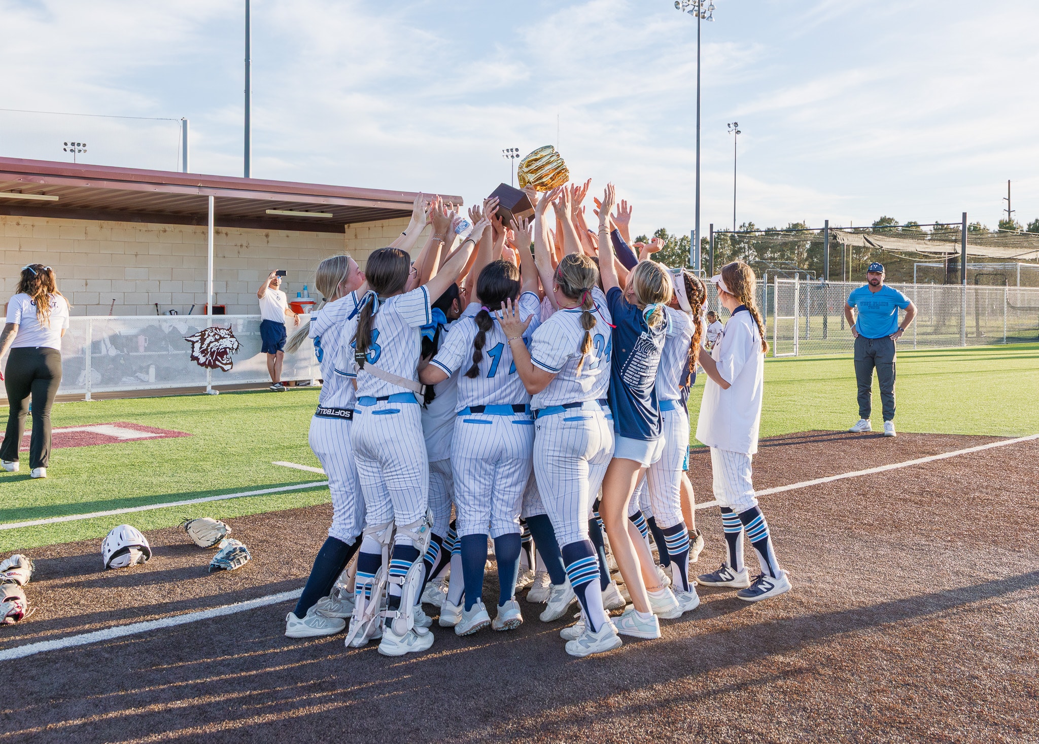 Girls softball team huddles and raises a trophy in celebration on the field with helmets on the ground nearby.