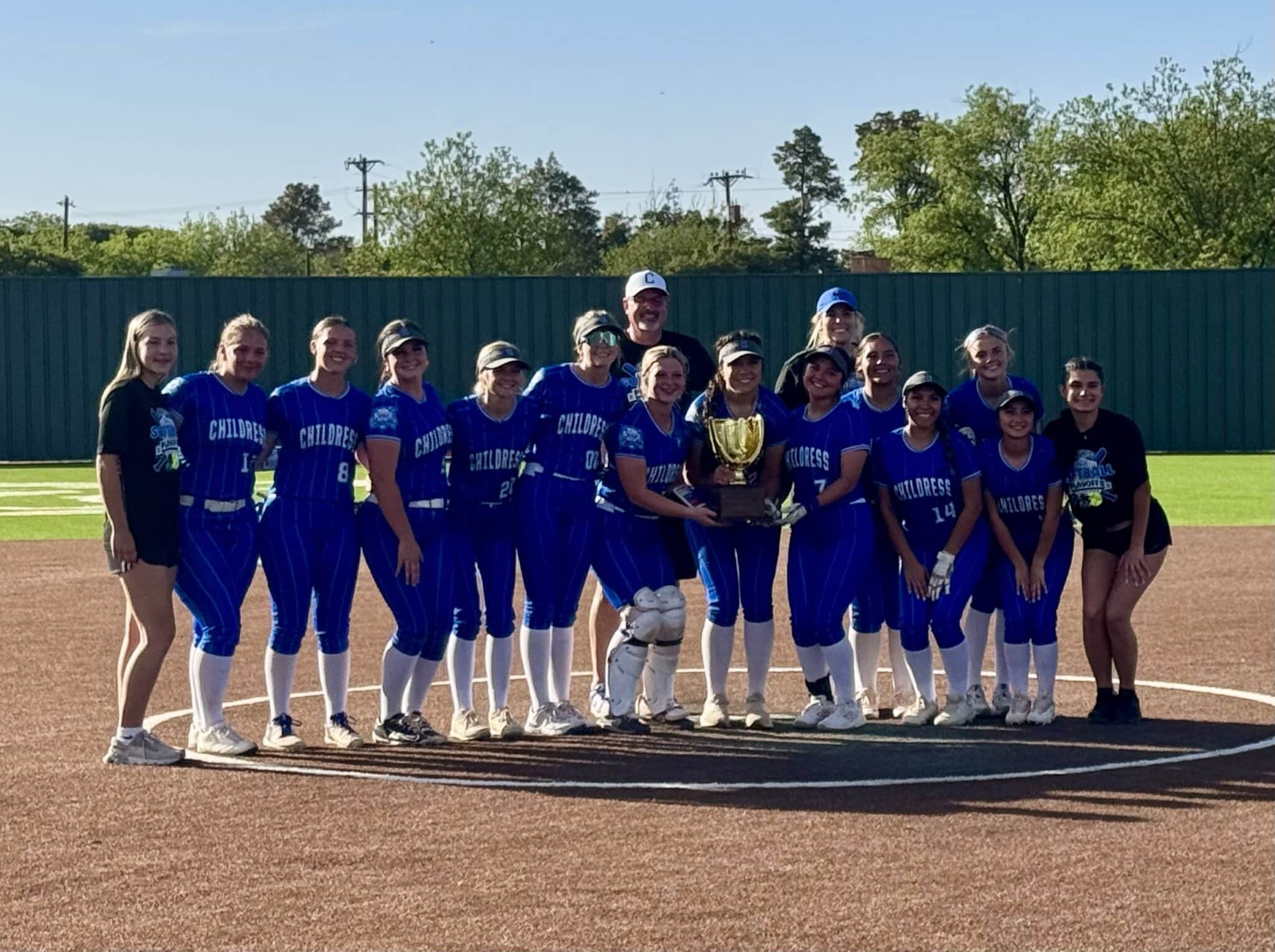 Girls softball team in blue uniforms posing with a trophy on a softball field in front of a green fence and blue sky.