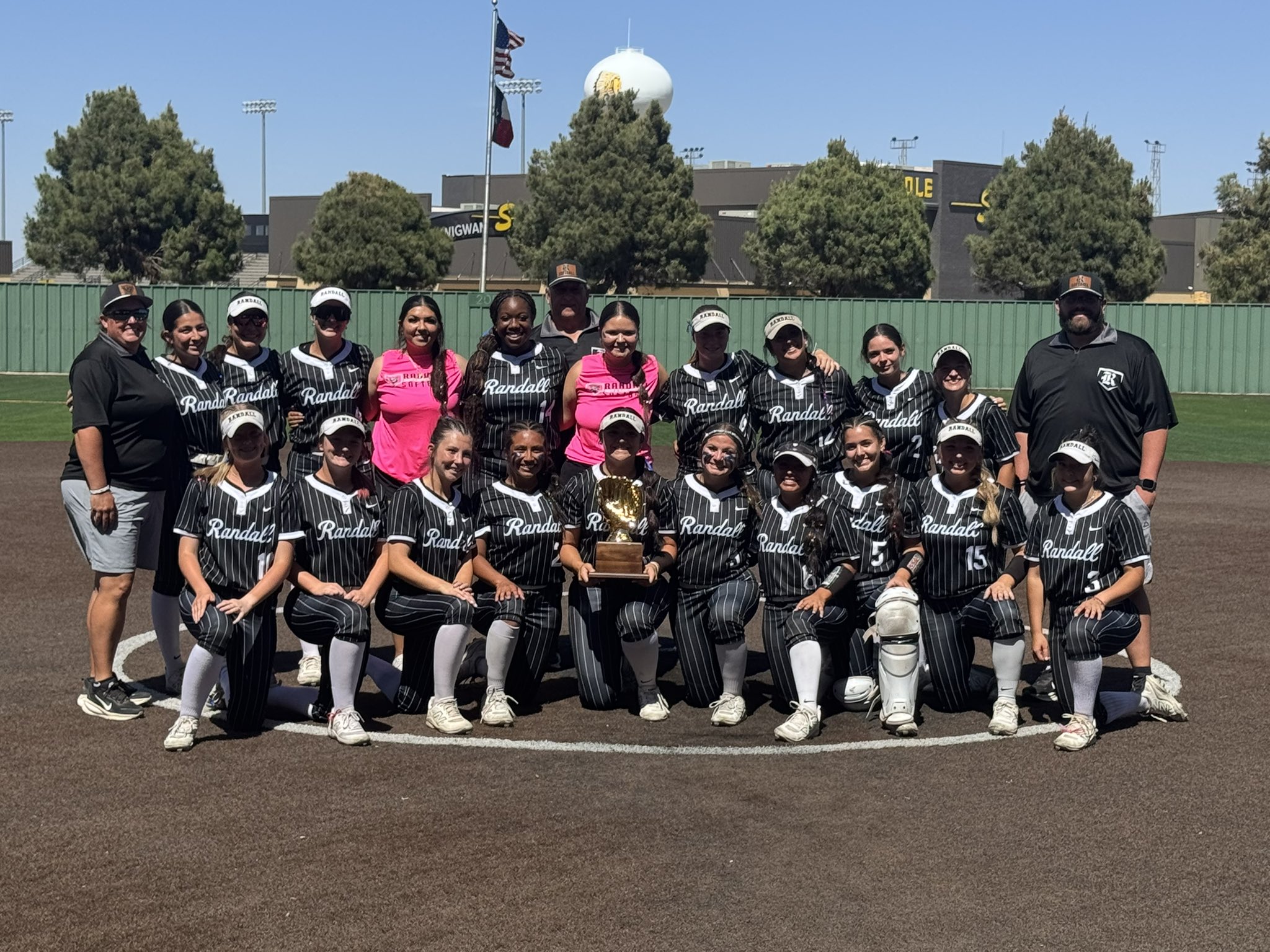 Group of female softball players in black pinstripe uniforms with 'Randall' written on jerseys, posing together on a baseball field with a trophy in the center; coaches at ends.