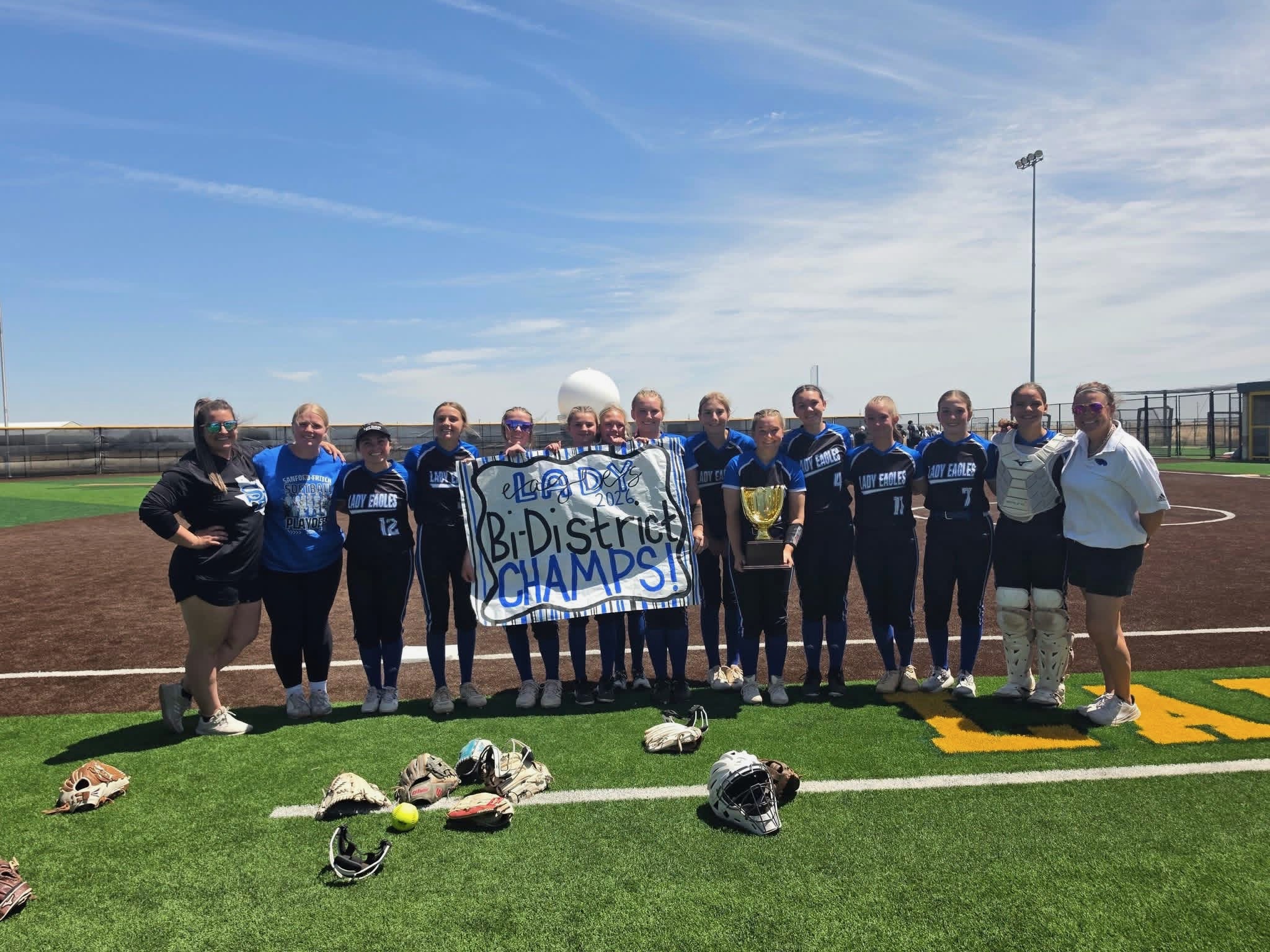Girls softball team posing on a field with a banner and trophy, gloves scattered on the turf under a clear blue sky