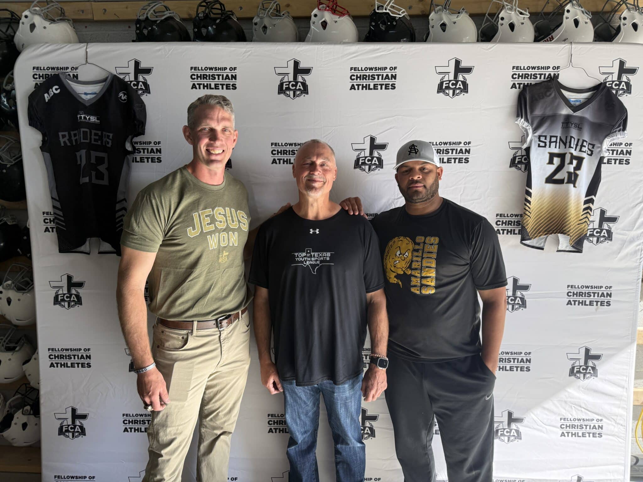 Three men pose together in front of a ChristianAthletes backdrop with hockey-style jerseys hanging behind them.