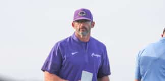 Man in a purple athletic shirt and cap holding a card, standing outdoors, facing the camera.