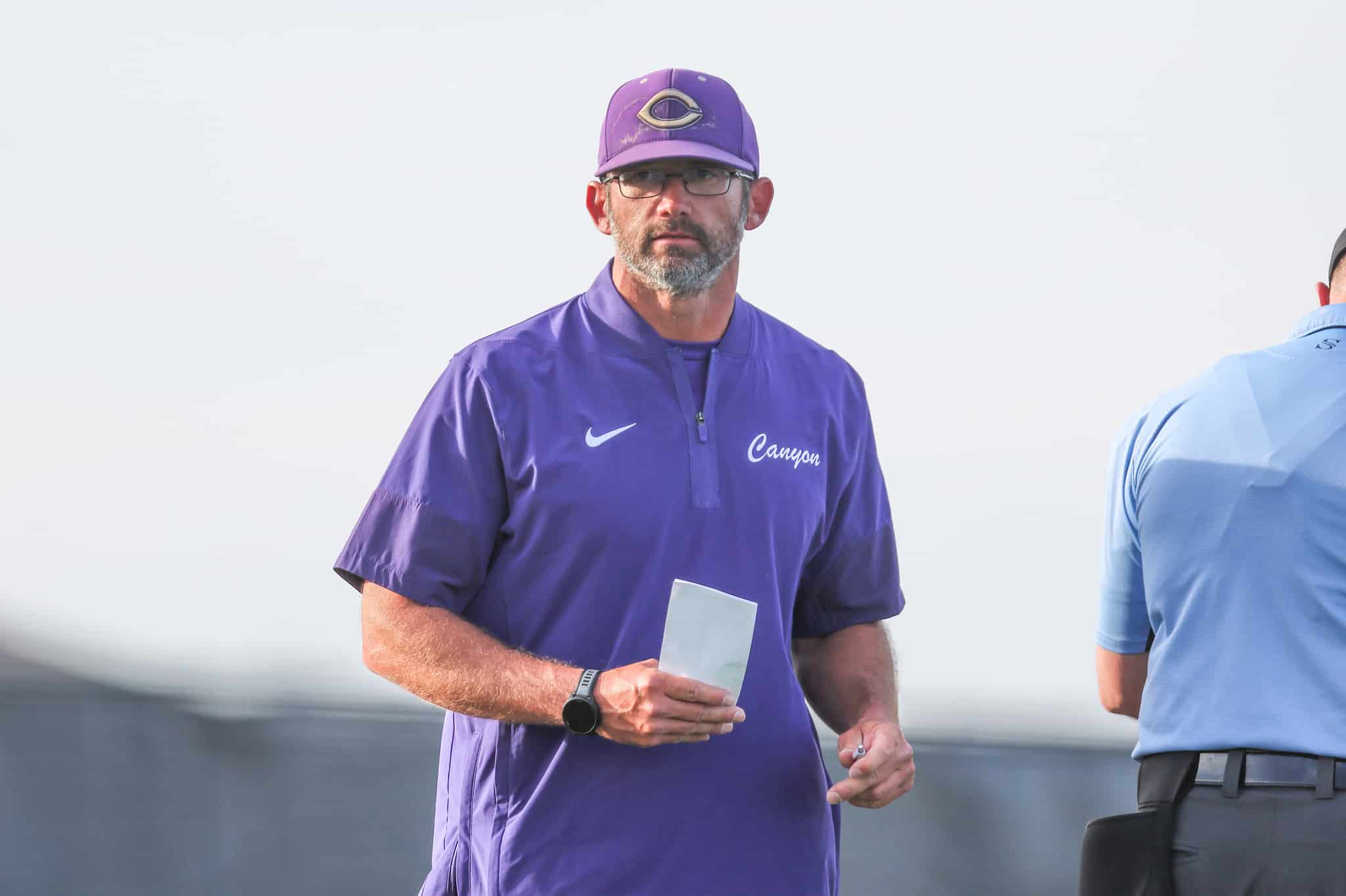 Man in a purple athletic shirt and cap holding a card, standing outdoors, facing the camera.