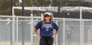 Athlete posing on a grassy field in a navy playoffs shirt, visor, sunglasses, and tattoos, with a chain-link fence behind her.