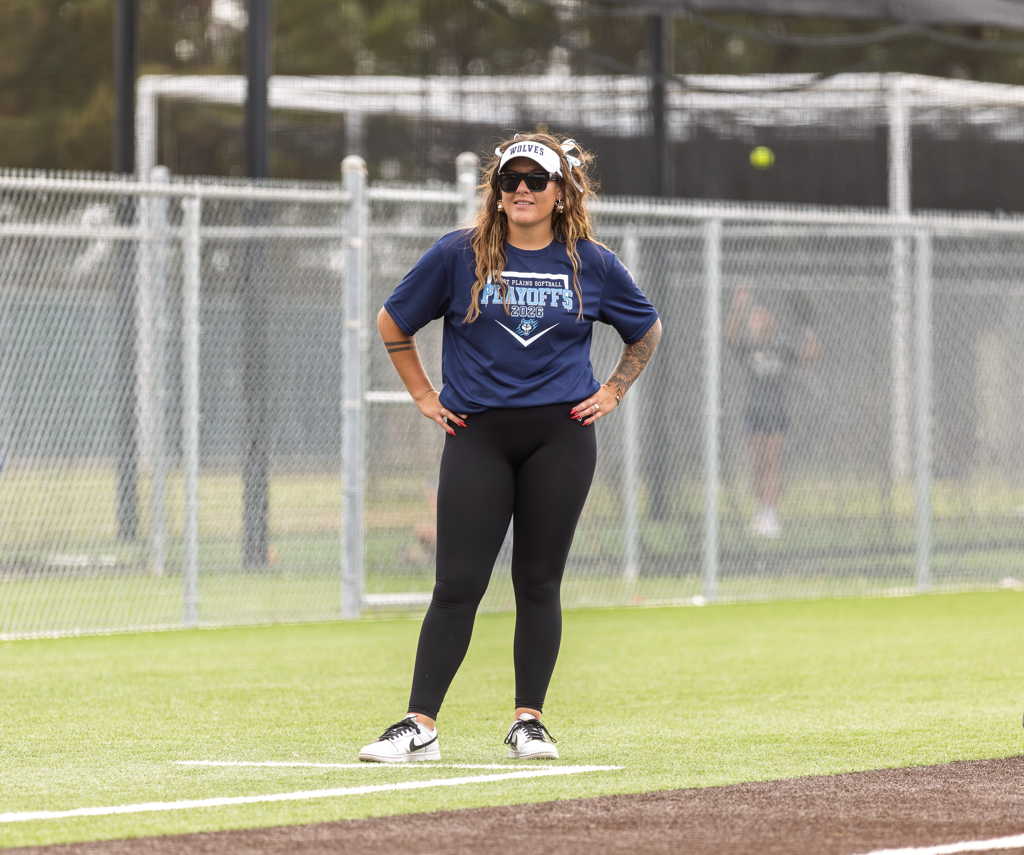 Athlete posing on a grassy field in a navy playoffs shirt, visor, sunglasses, and tattoos, with a chain-link fence behind her.
