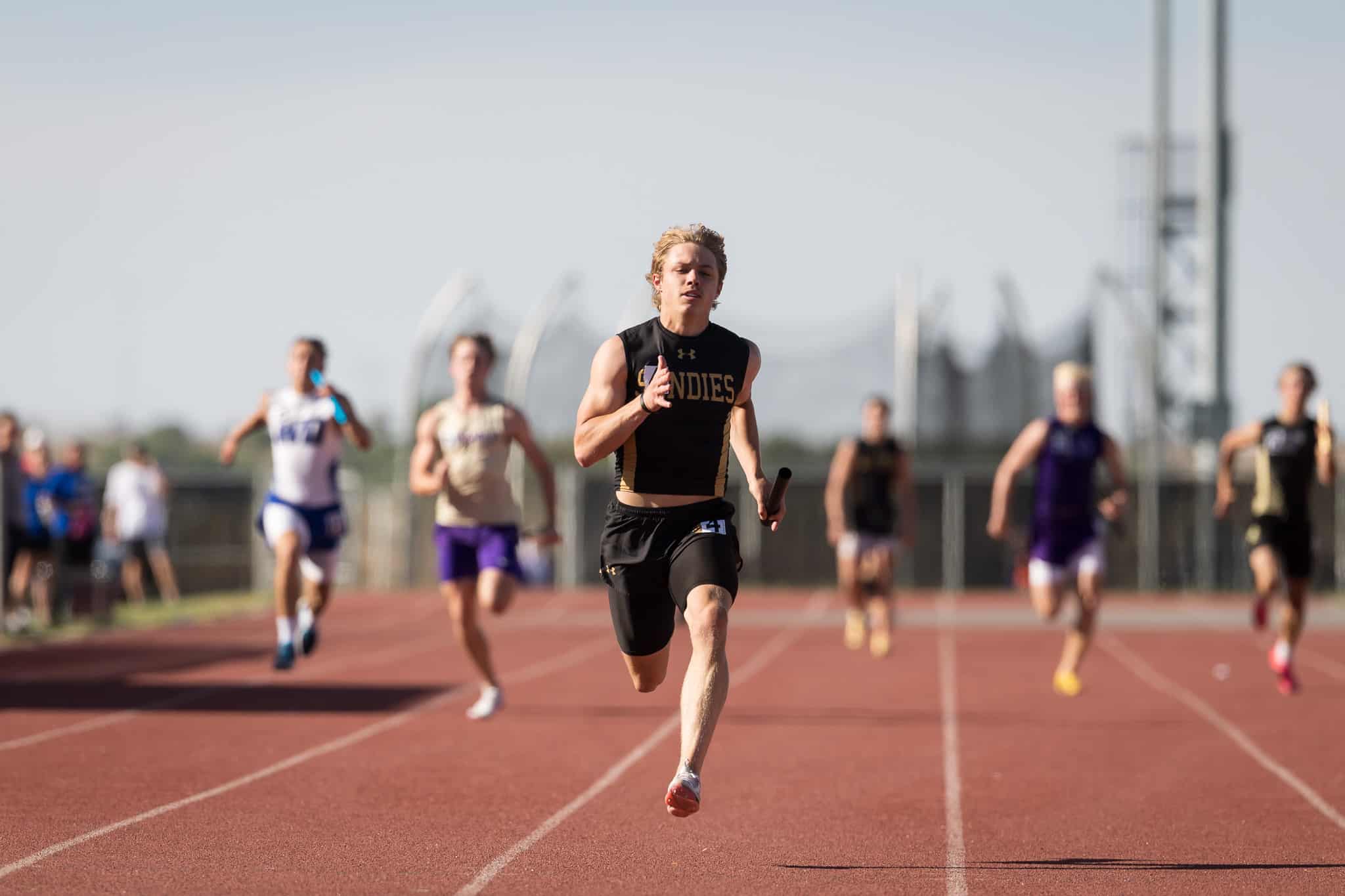 Male sprinter in a black singlet leads a track race on a red running track with competitors blurred in the background.
