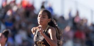 Female runner in a black and gold outfit mid-race, with a blurred crowd in the background.