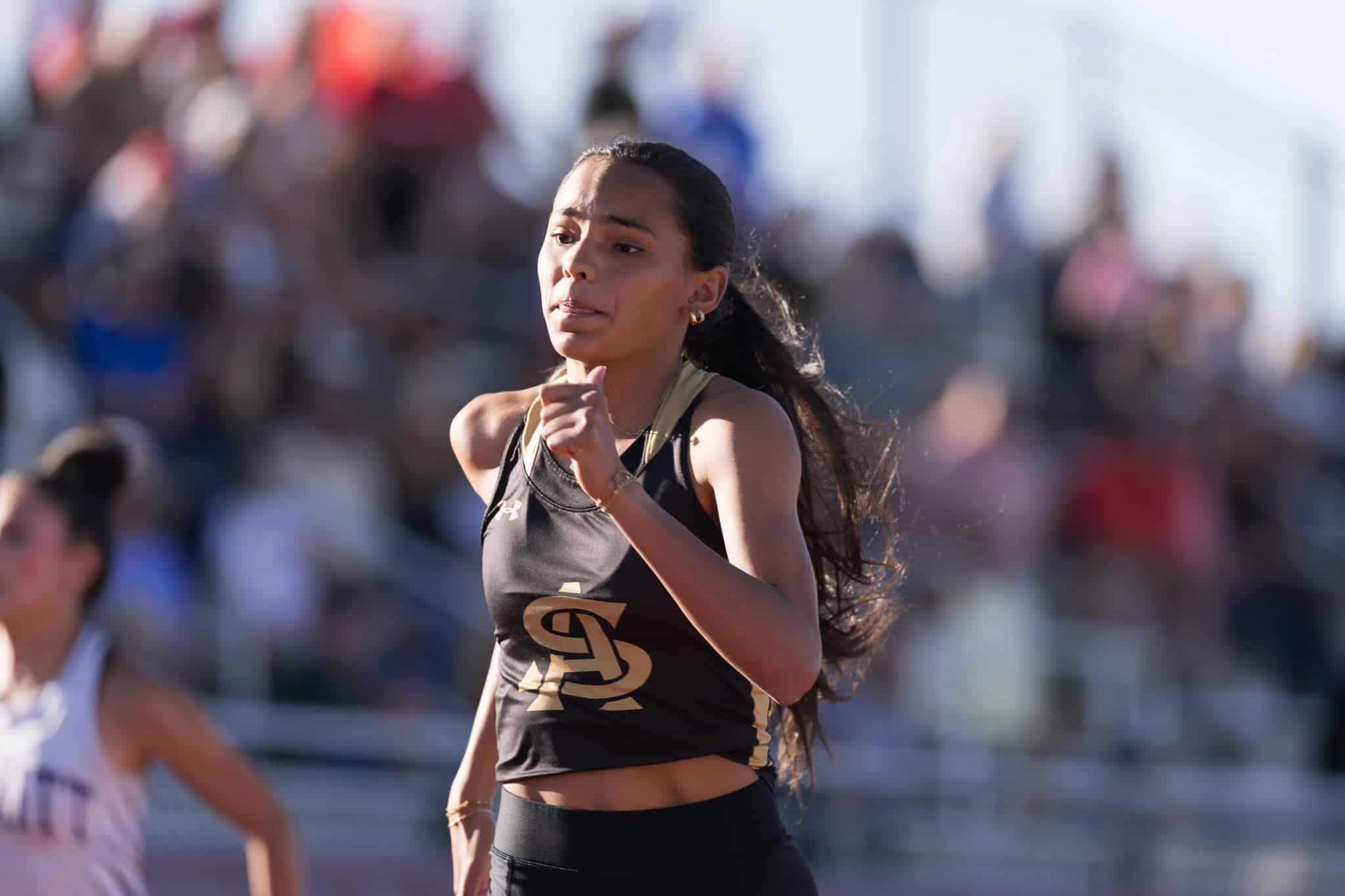 Female runner in a black and gold outfit mid-race, with a blurred crowd in the background.