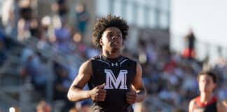 Male sprinter in a black sleeveless top with a white 'M' logo, running toward the camera at a crowded outdoor track meet.