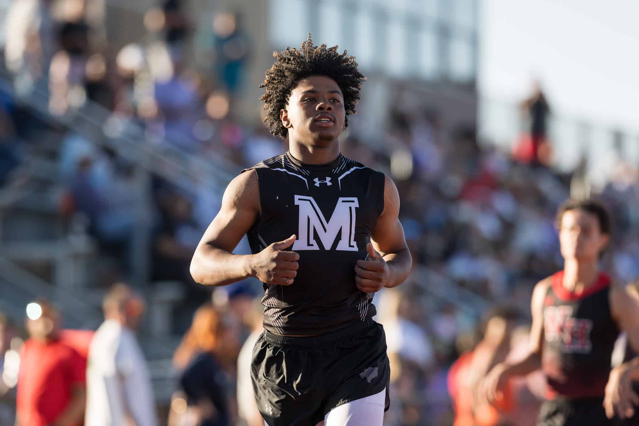 Male sprinter in a black sleeveless top with a white 'M' logo, running toward the camera at a crowded outdoor track meet.