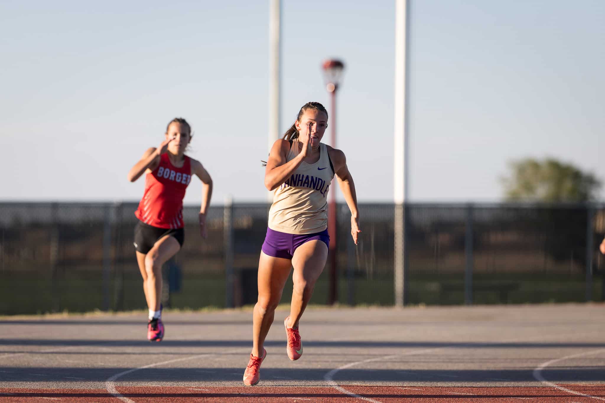 Two female sprinters race down an outdoor track, foreground runner in tan top and purple shorts leading as a blurred rival follows behind.