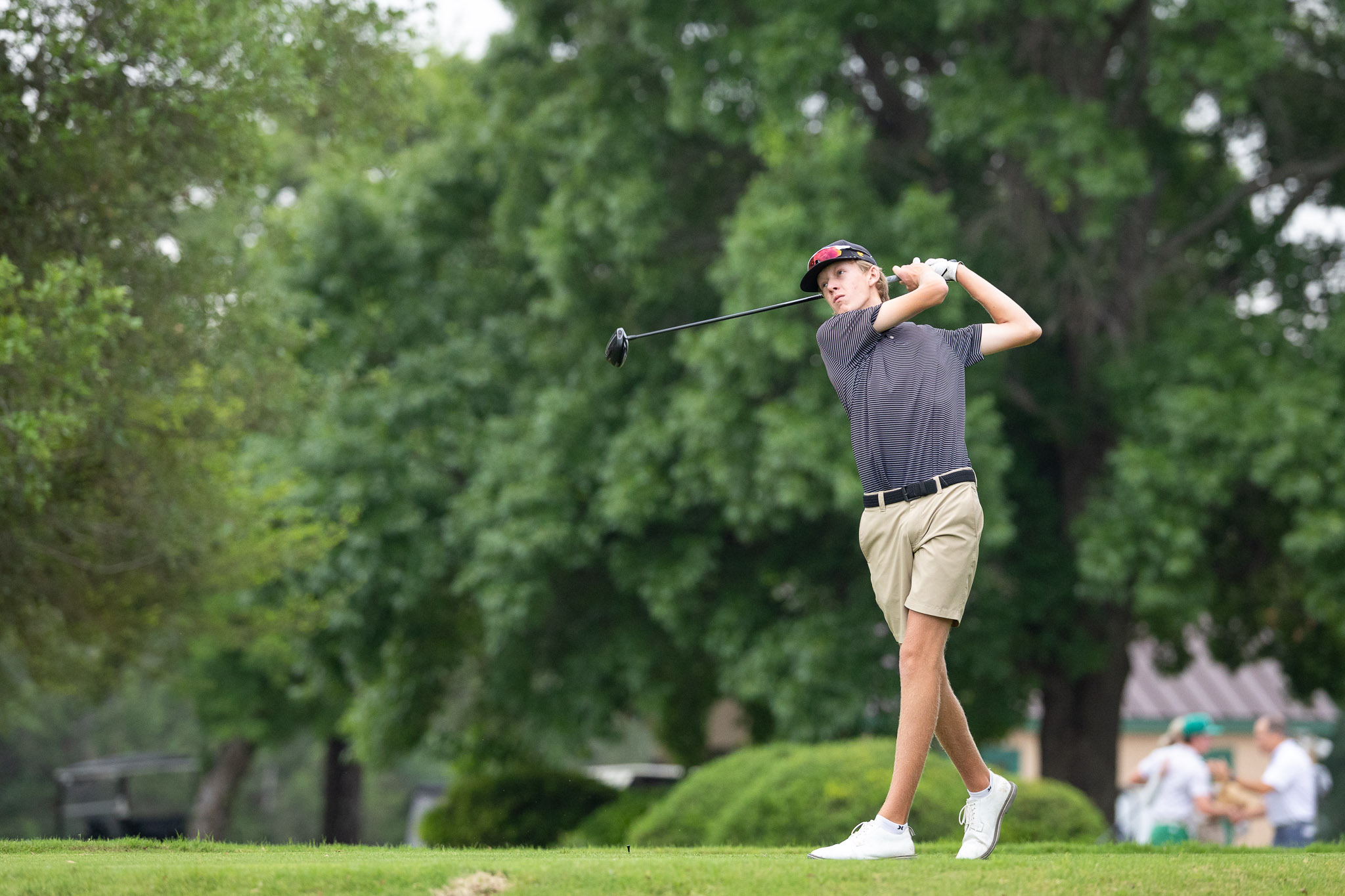 Young man in a striped shirt and cap mid-swing with a golf club on a green golf course.