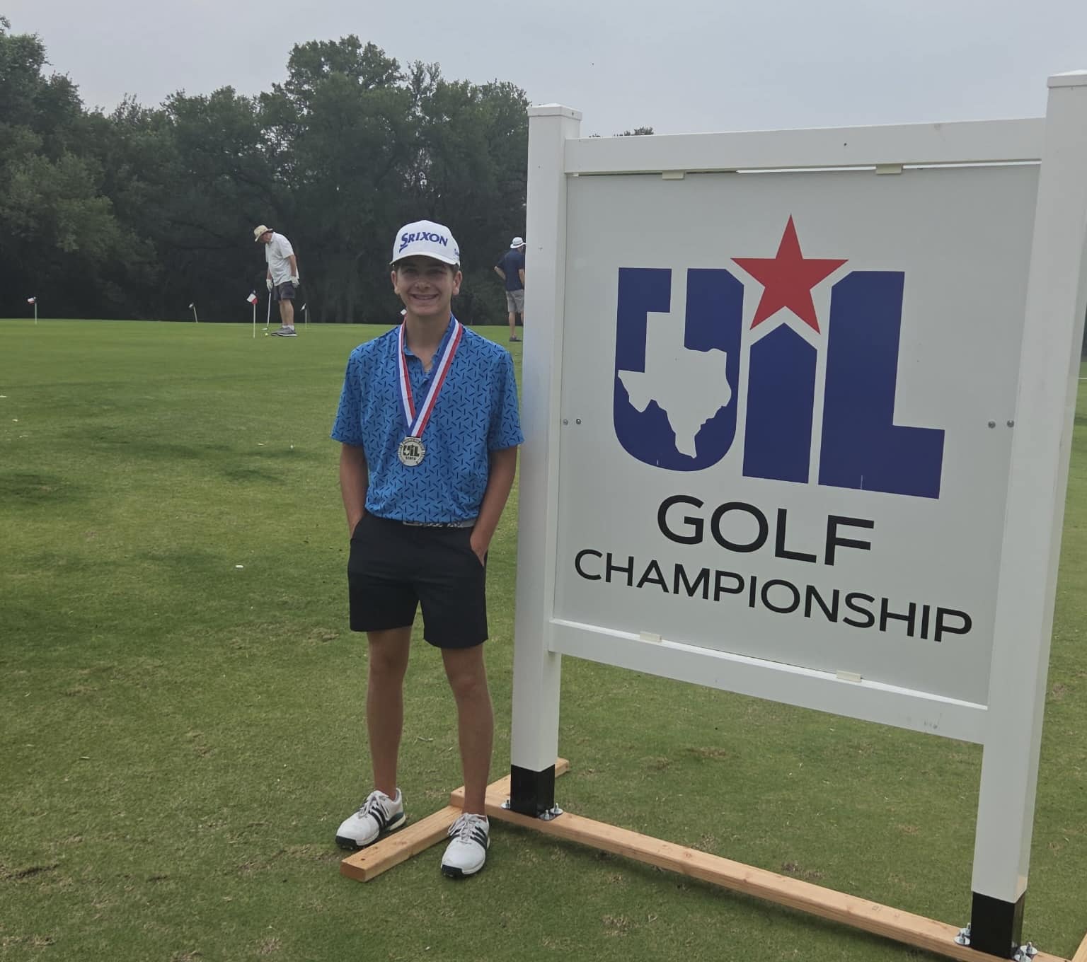 Young golfer wearing a blue patterned shirt and white cap with a medal stands beside a UIL Golf Championship sign on a green golf course.