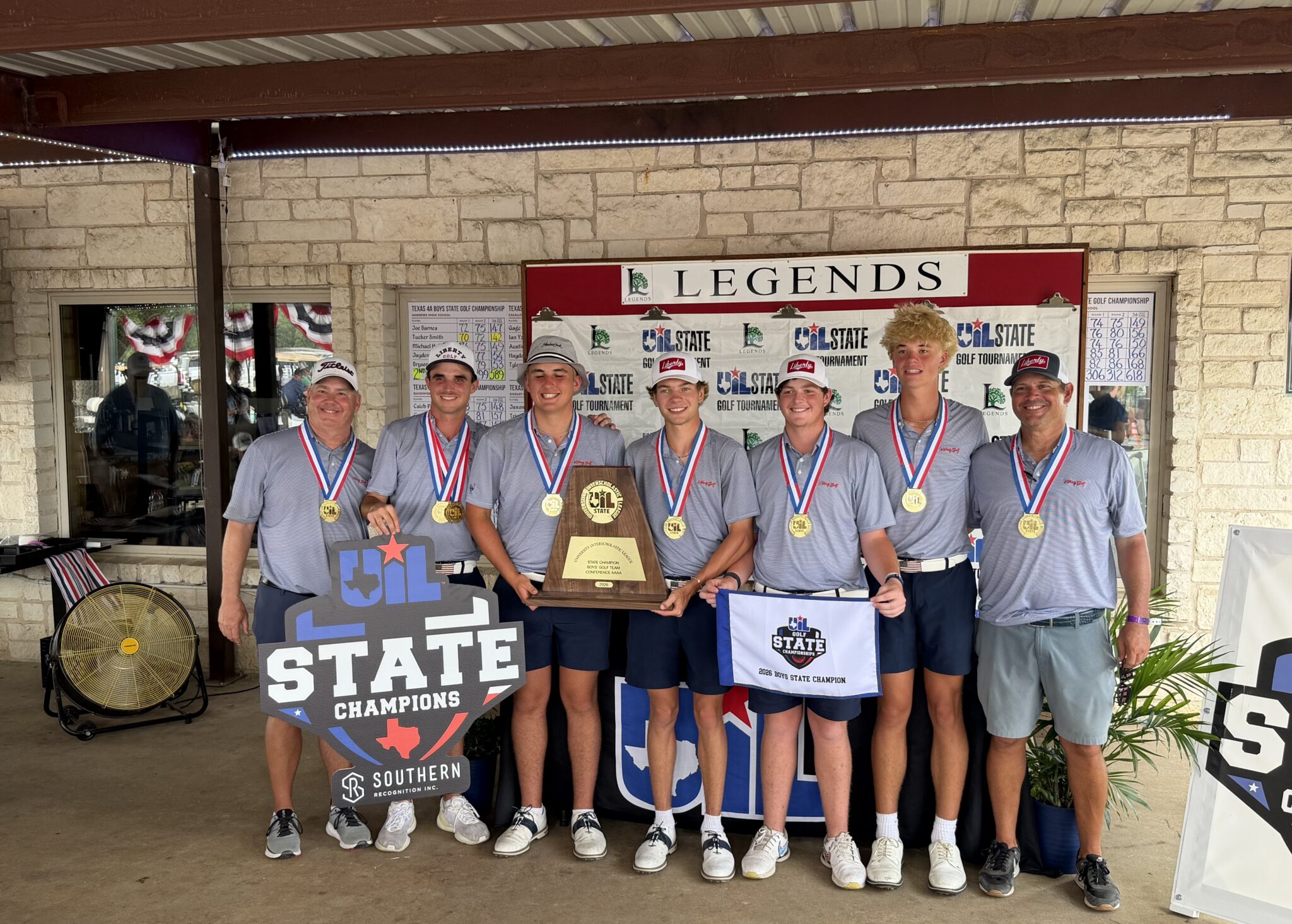 Seven young golfers in gray shirts with gold medals pose together in front of a Legends State Championships banner, holding a large trophy and banners.