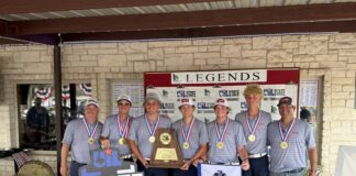 Seven young golfers in gray shirts with gold medals pose together in front of a Legends State Championships banner, holding a large trophy and banners.