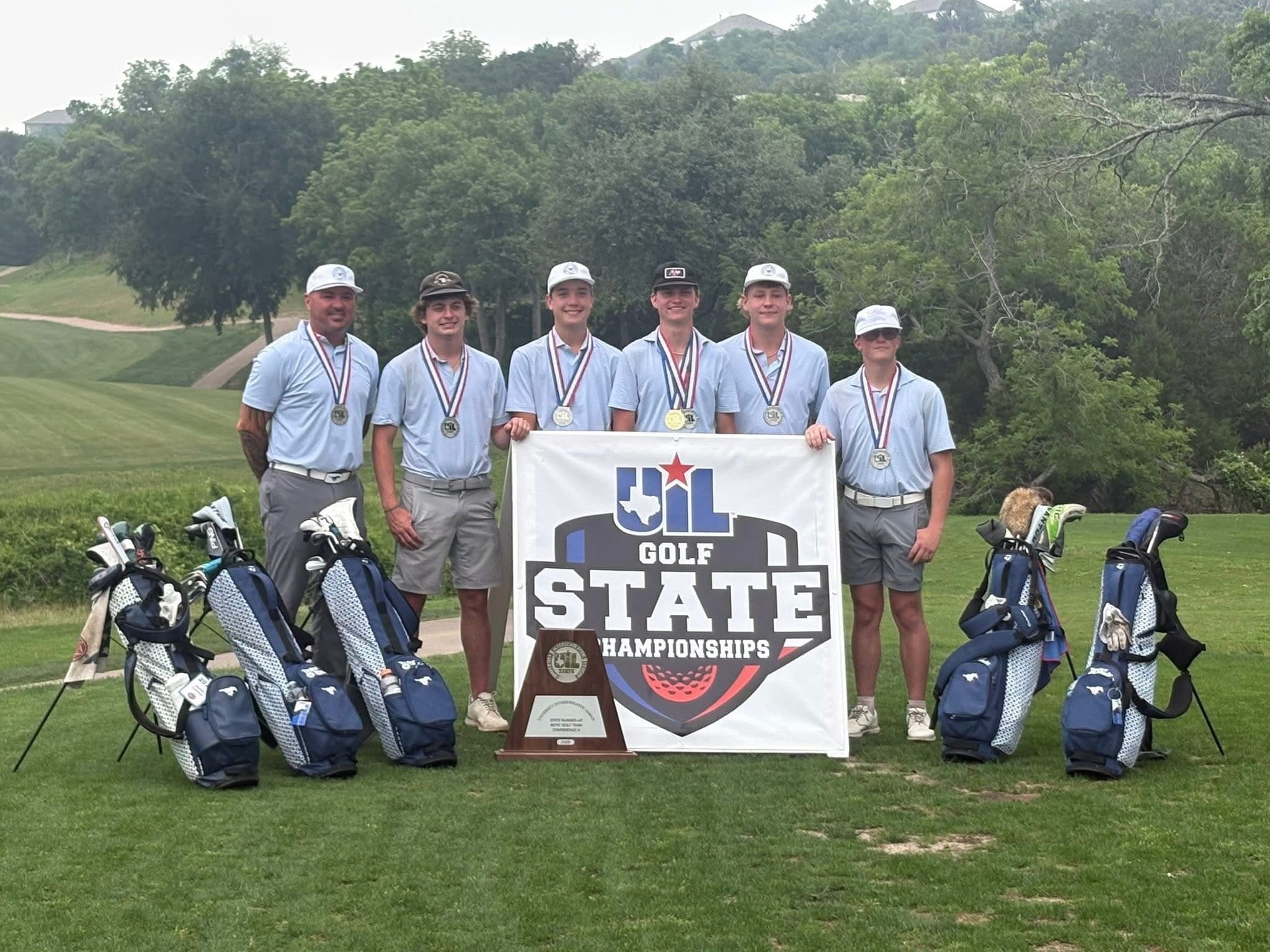 Six golfers in light blue shirts and caps pose with medals on a golf course, holding a banner that reads 'GOLF STATE CHAMPIONSHIPS' with a trophy in front.