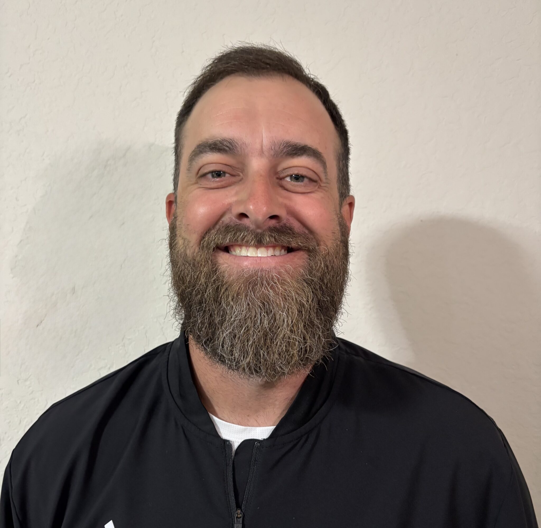 Smiling man with a beard and short dark hair wearing a black jacket, against a light textured wall.
