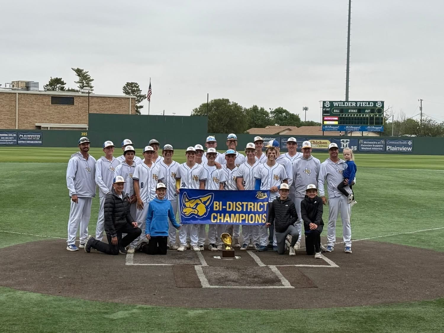 Youth baseball team posing on the field with a Bi-District Champions banner and trophy in front of them on home plate.