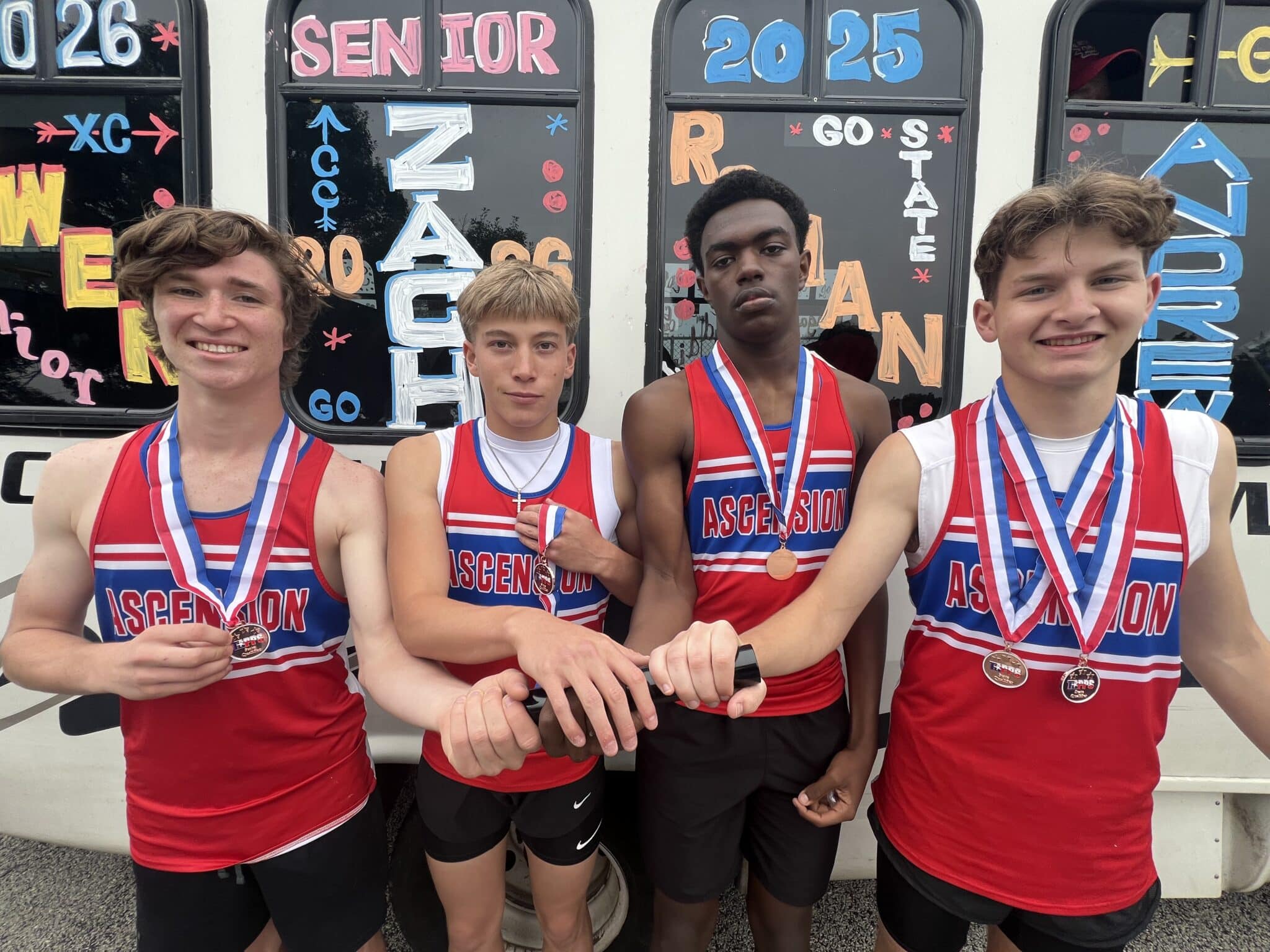 Four male runners in red, white, and blue Ascension uniforms pose with medals around their necks in front of a bus covered in colorful chalk art and numbers.