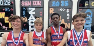 Four male runners in red, white, and blue Ascension uniforms pose with medals around their necks in front of a bus covered in colorful chalk art and numbers.
