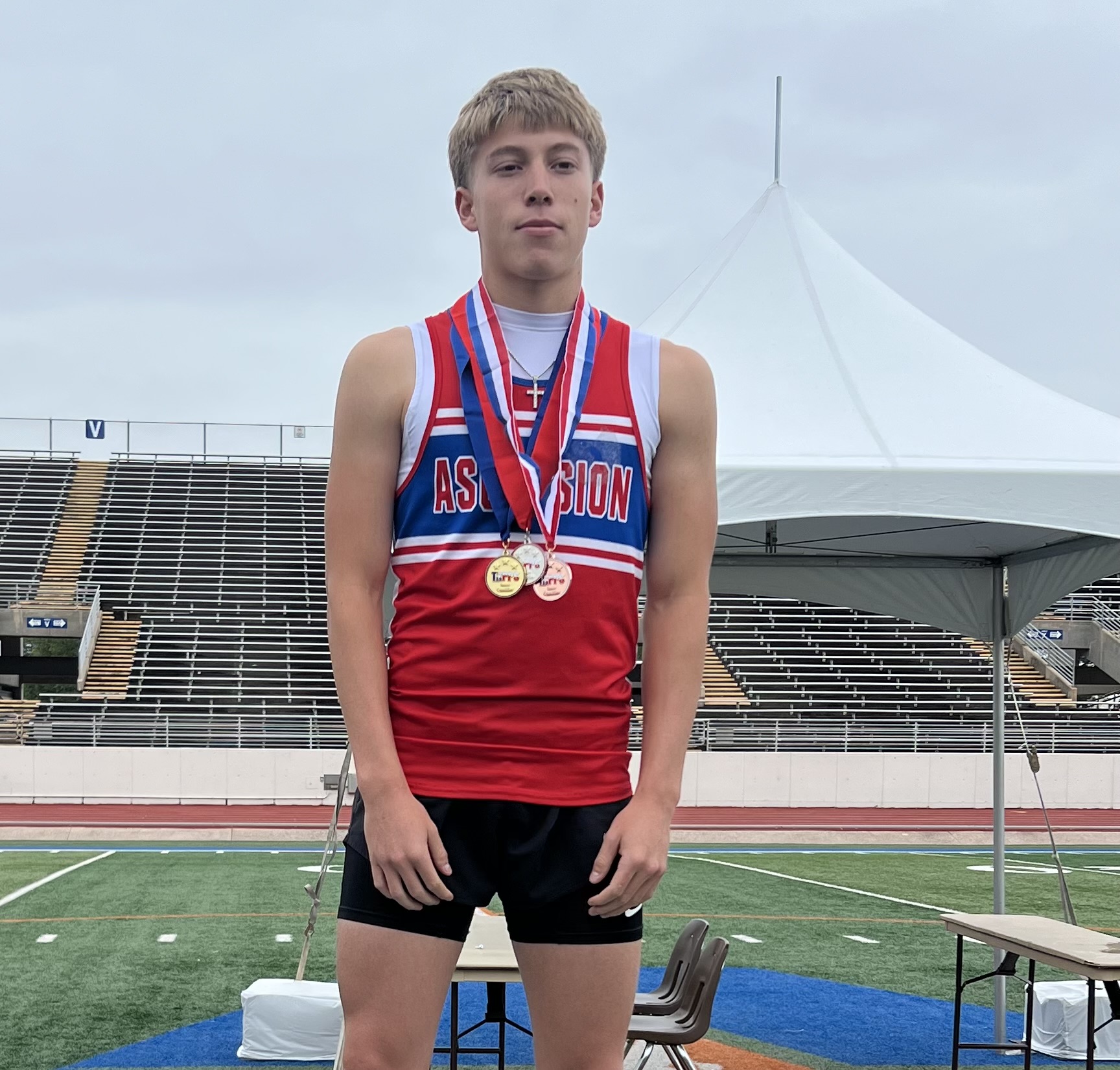 Young male athlete in a red and blue track uniform with multiple medals around his neck, standing on a track with bleachers behind him and a white tent in the background.