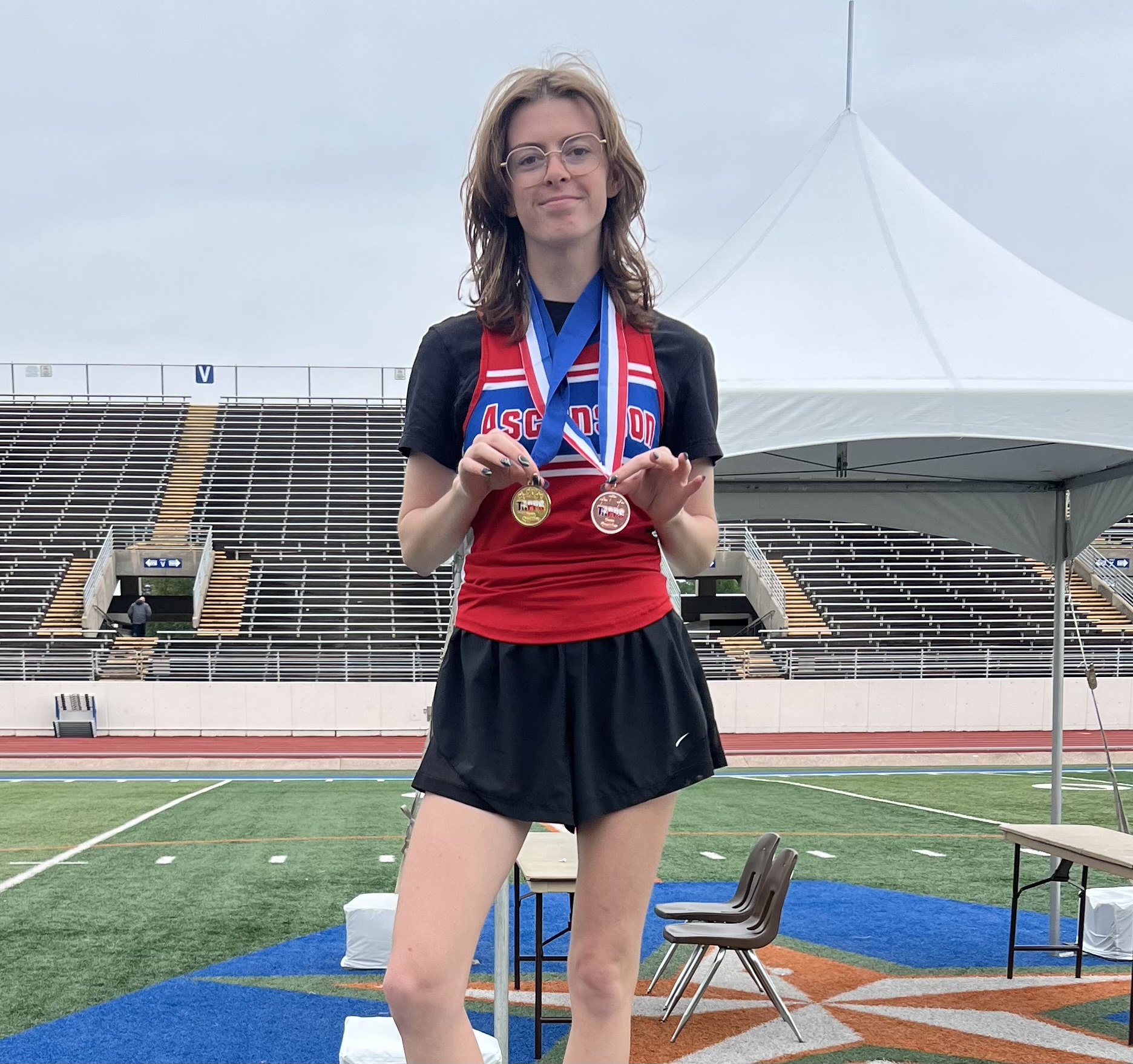 Young female athlete with two medals around her neck on a track, bleachers behind and a white tent nearby.