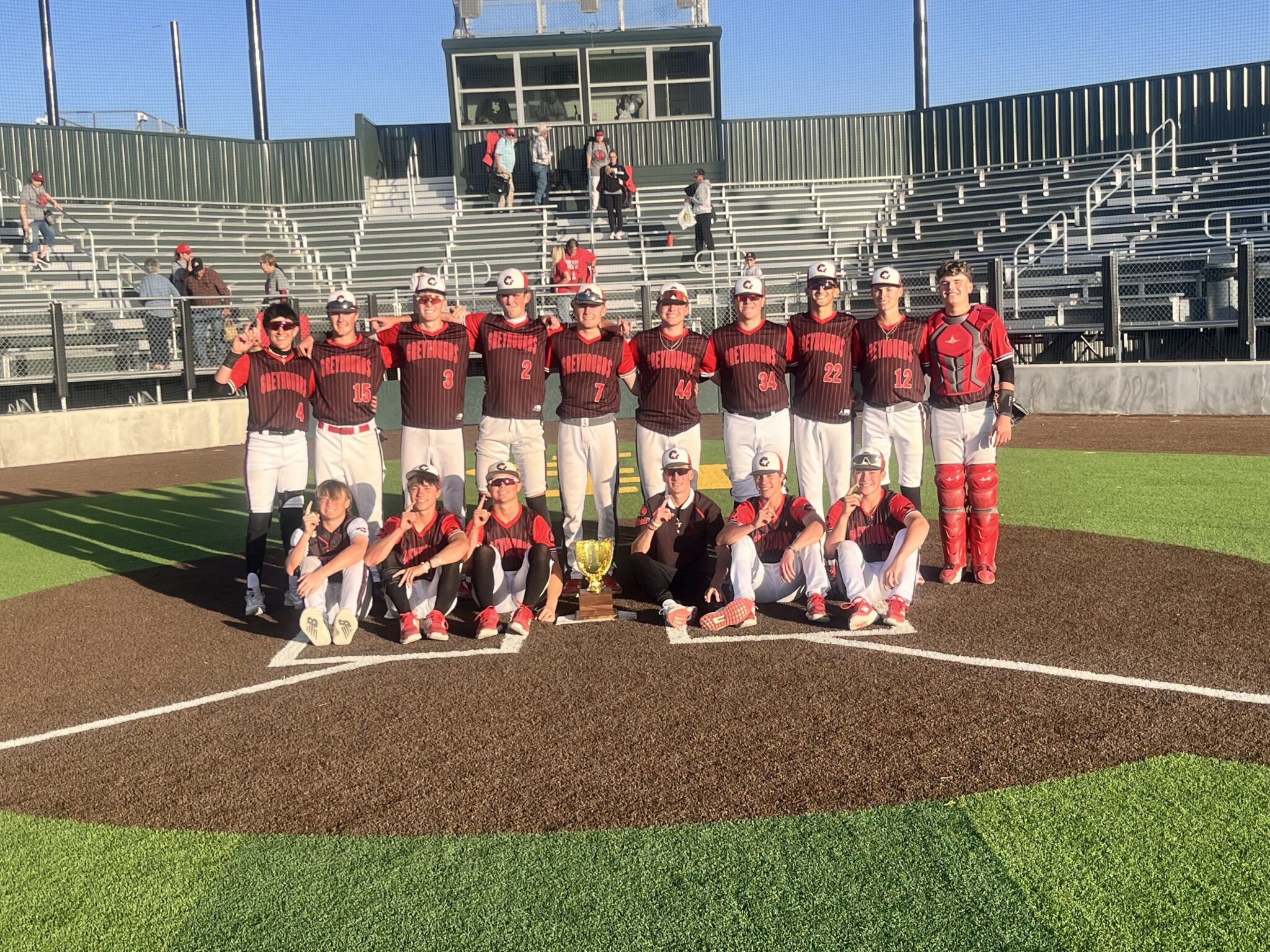 A baseball team wearing black and red uniforms poses together on the field with a trophy in front of them, smiling at the camera.