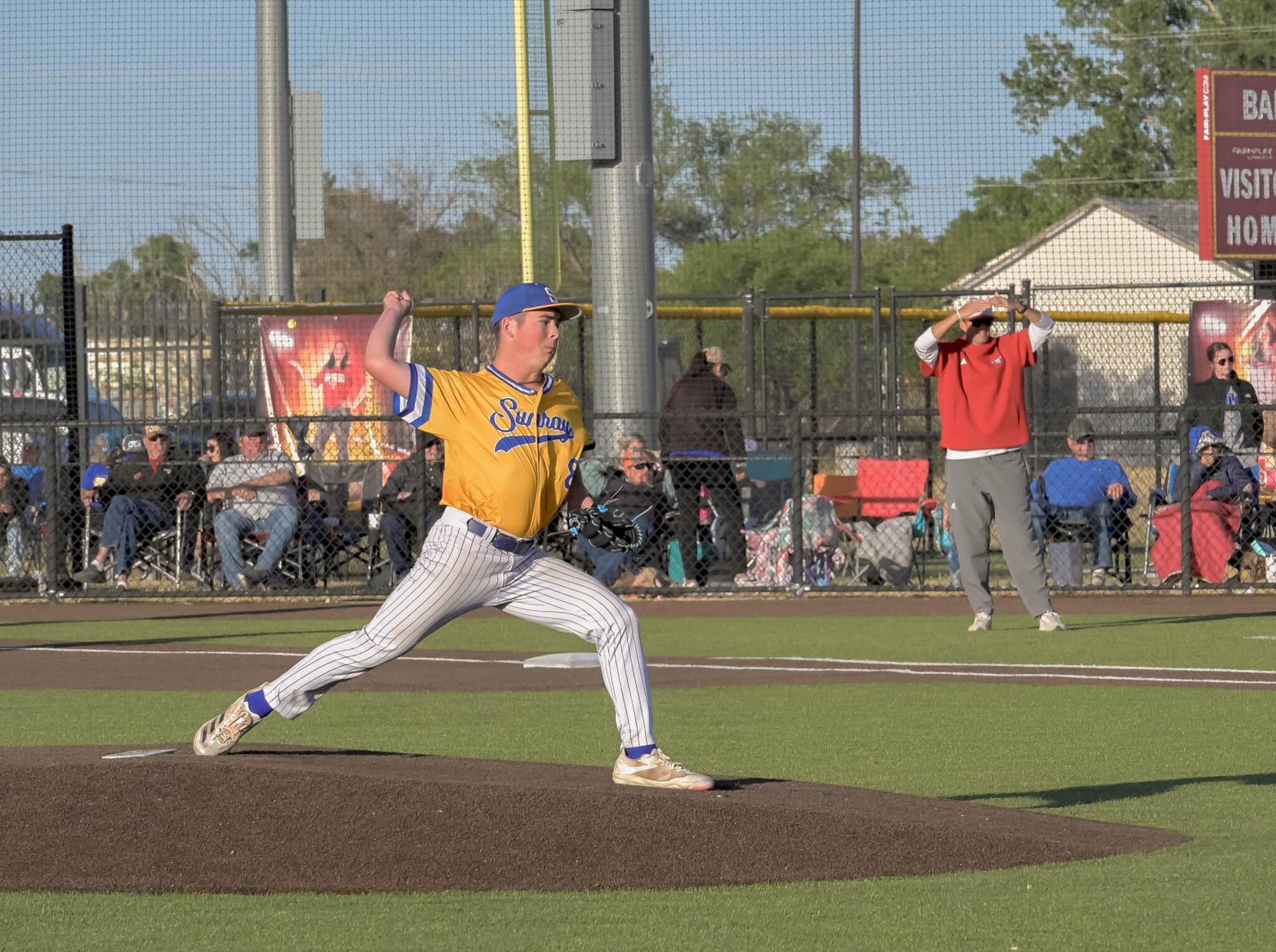 Baseball pitcher in yellow jersey delivering a pitch on the mound with spectators behind a chain-link fence.