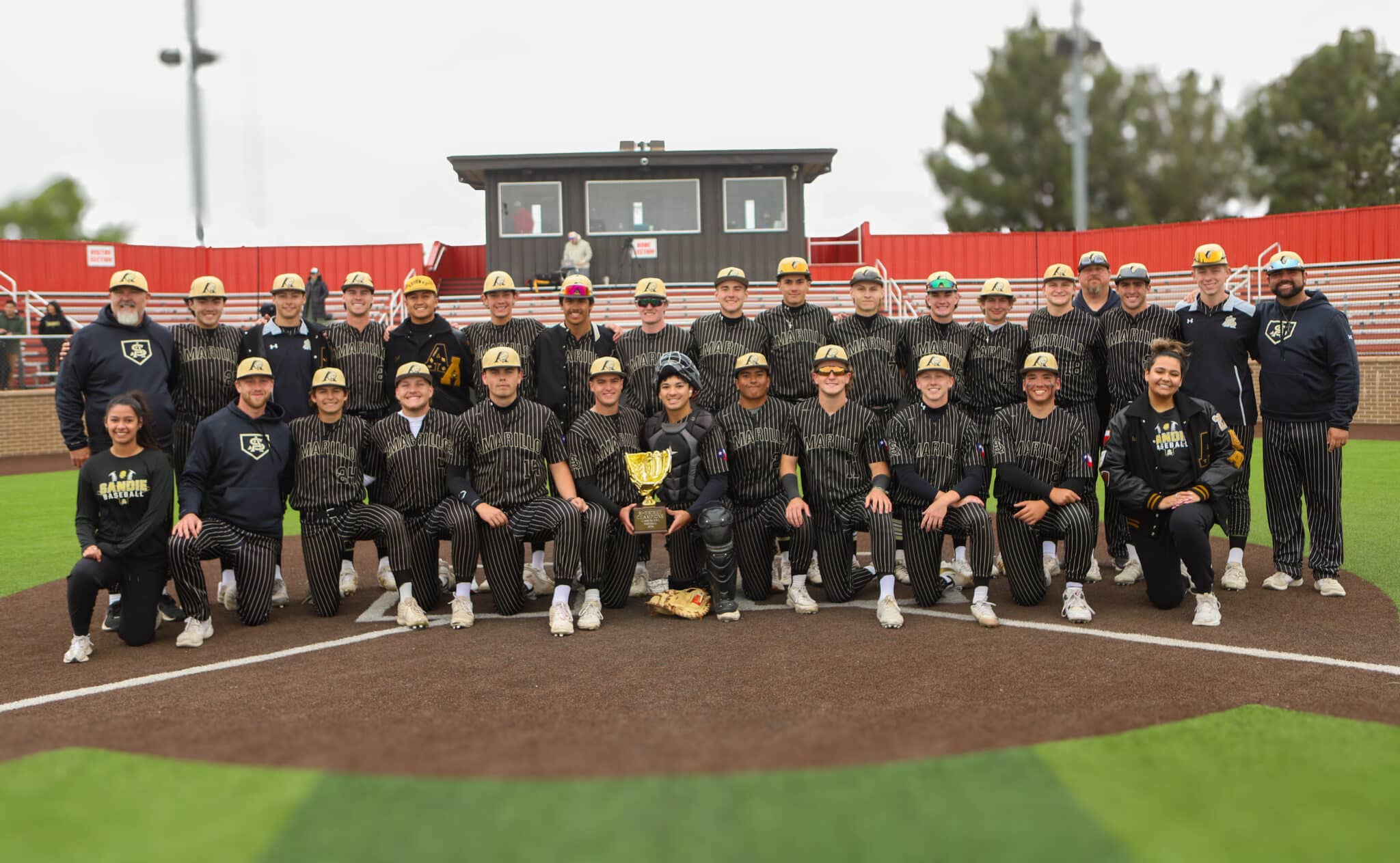 Baseball team posing for a group photo on the field with a trophy at center.