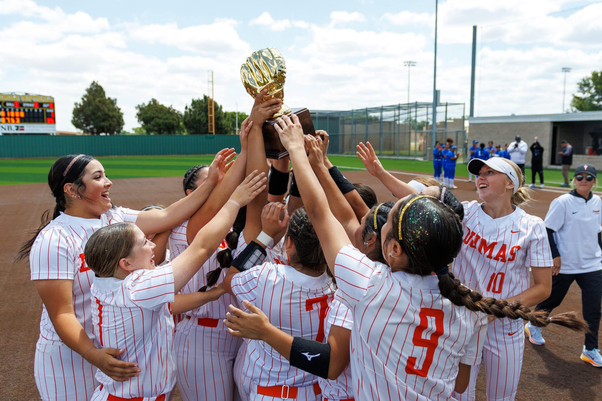 IMG_2052 | Press Pass Sports Girls softball team lifts a gold trophy in celebration on the field, wearing white uniforms with red pinstripes.