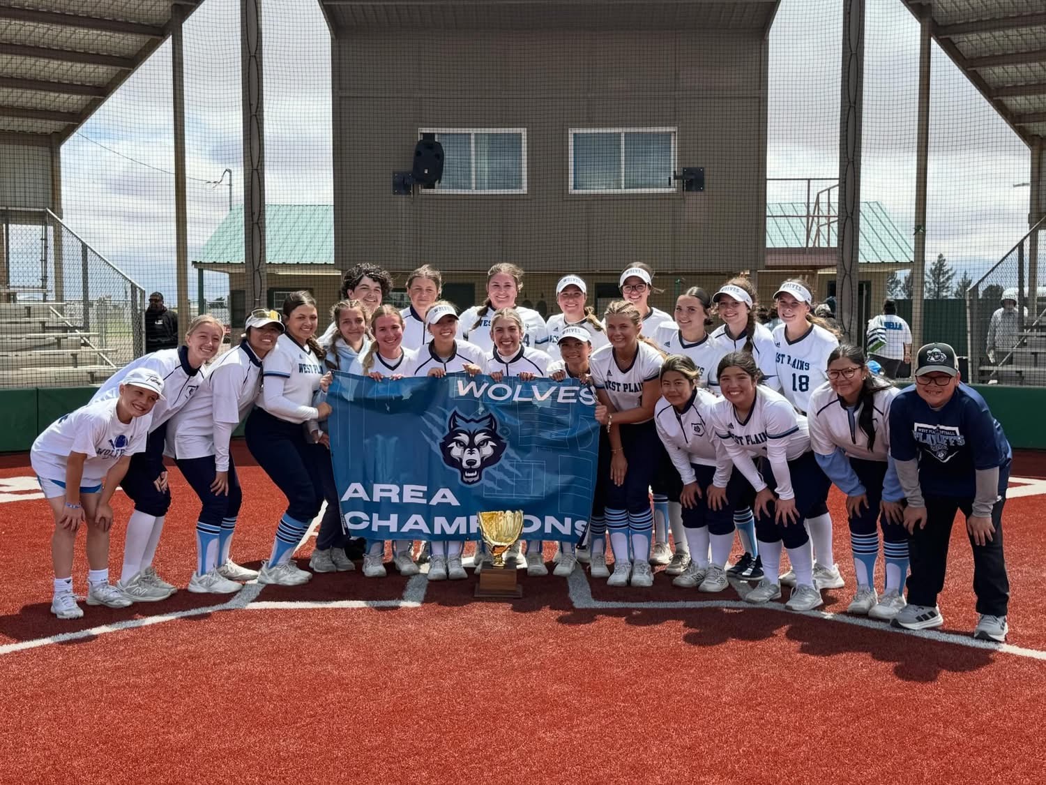 Girls softball team posing on a red clay field, holding a blue banner that reads 'WOLVES AREA CHAMPIONS' with a trophy in front.