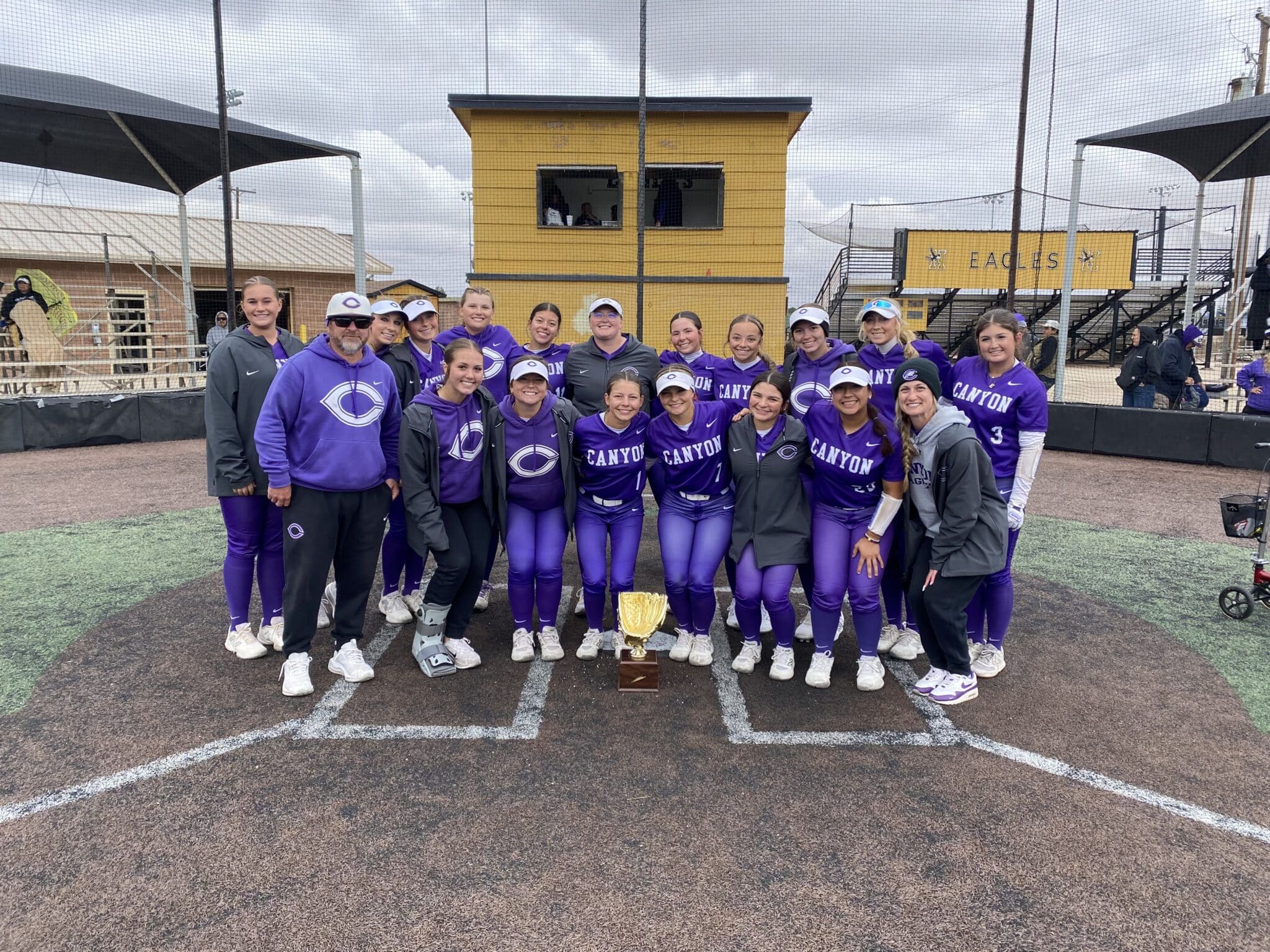 IMG_3728 | Press Pass Sports Group of female softball players in purple uniforms posing with a trophy on a baseball diamond.
