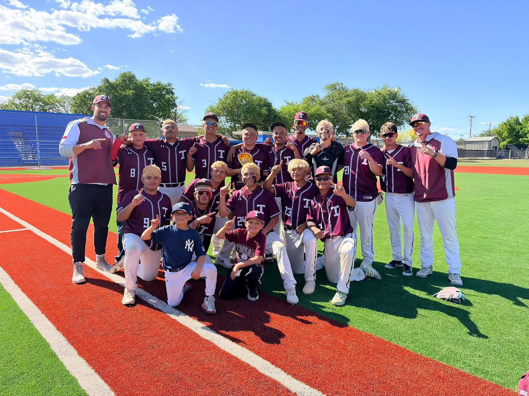 Group of young baseball players in maroon uniforms posing with coaches on a sunny field after a game.