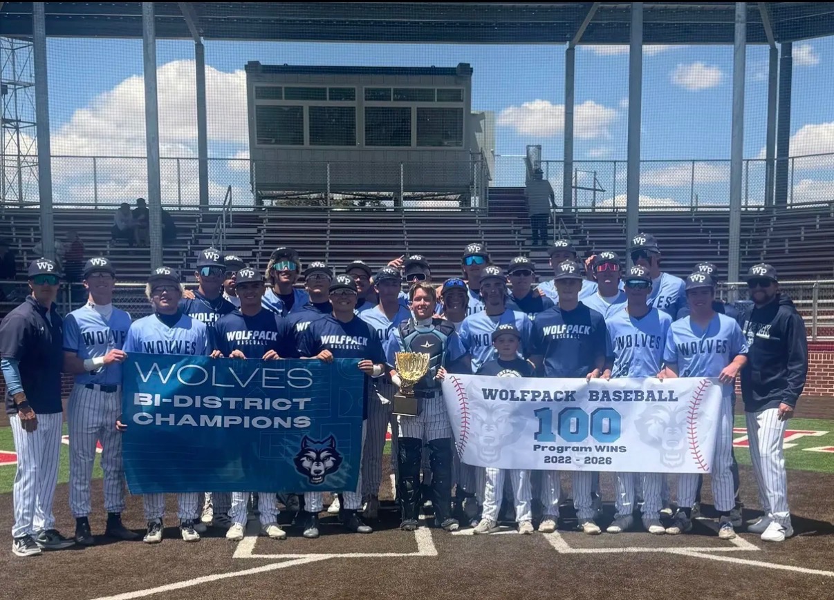 Group of Wolfpack/Wolves baseball players in blue uniforms pose with a large trophy and two banners on the field, celebrating bi-district champions.