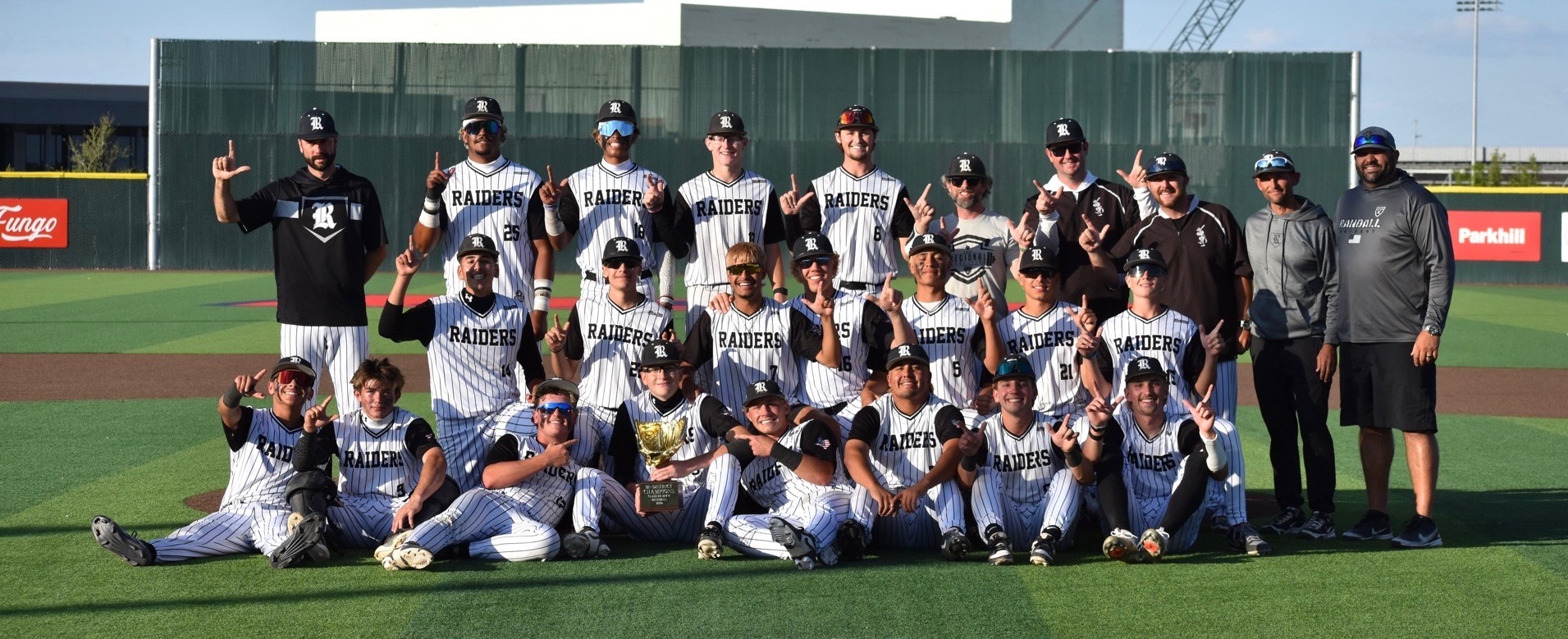 Raiders baseball team posing on the field with a trophy after a victory, group photo with players and coaches guardrails none